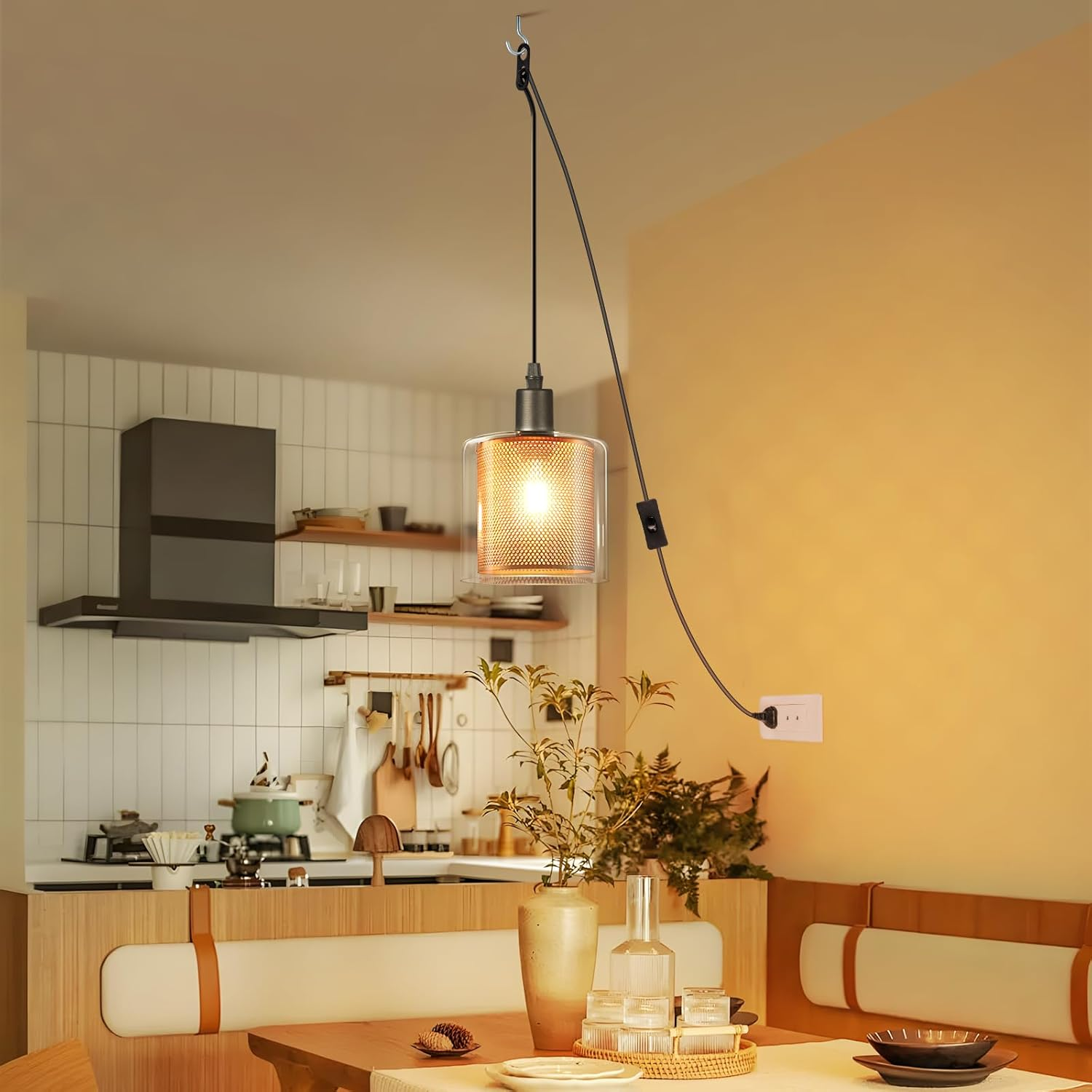 Una lampada a sospensione illuminata è appesa sopra un tavolo. Cucina con fornello, cappa e zona pranzo. Toni beige e arancioni.