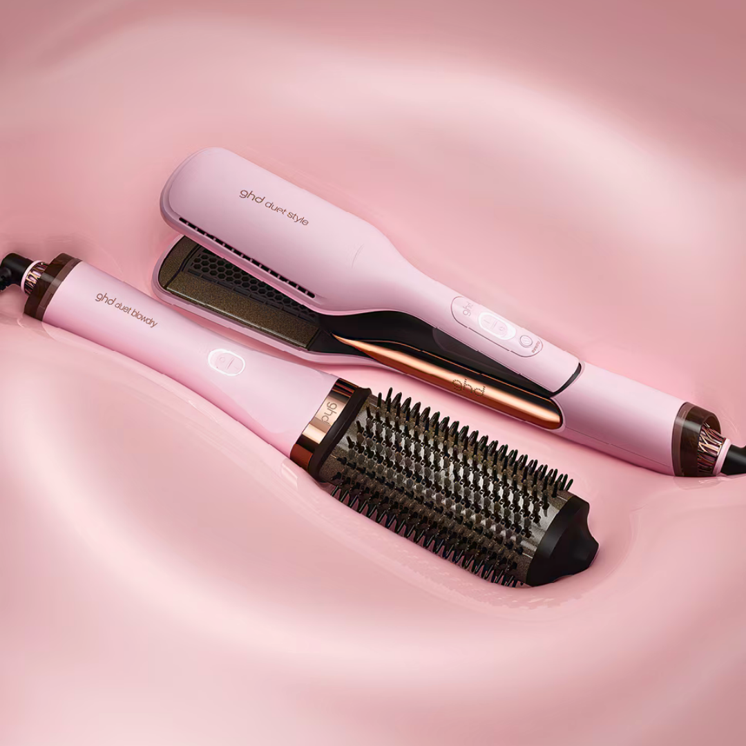 Zwei rosa GHD-Haarstyling-Tools auf rosa Oberfläche. Eines ist eine Bürste, das andere ein Glätteisen. Beide haben metallische Akzente.