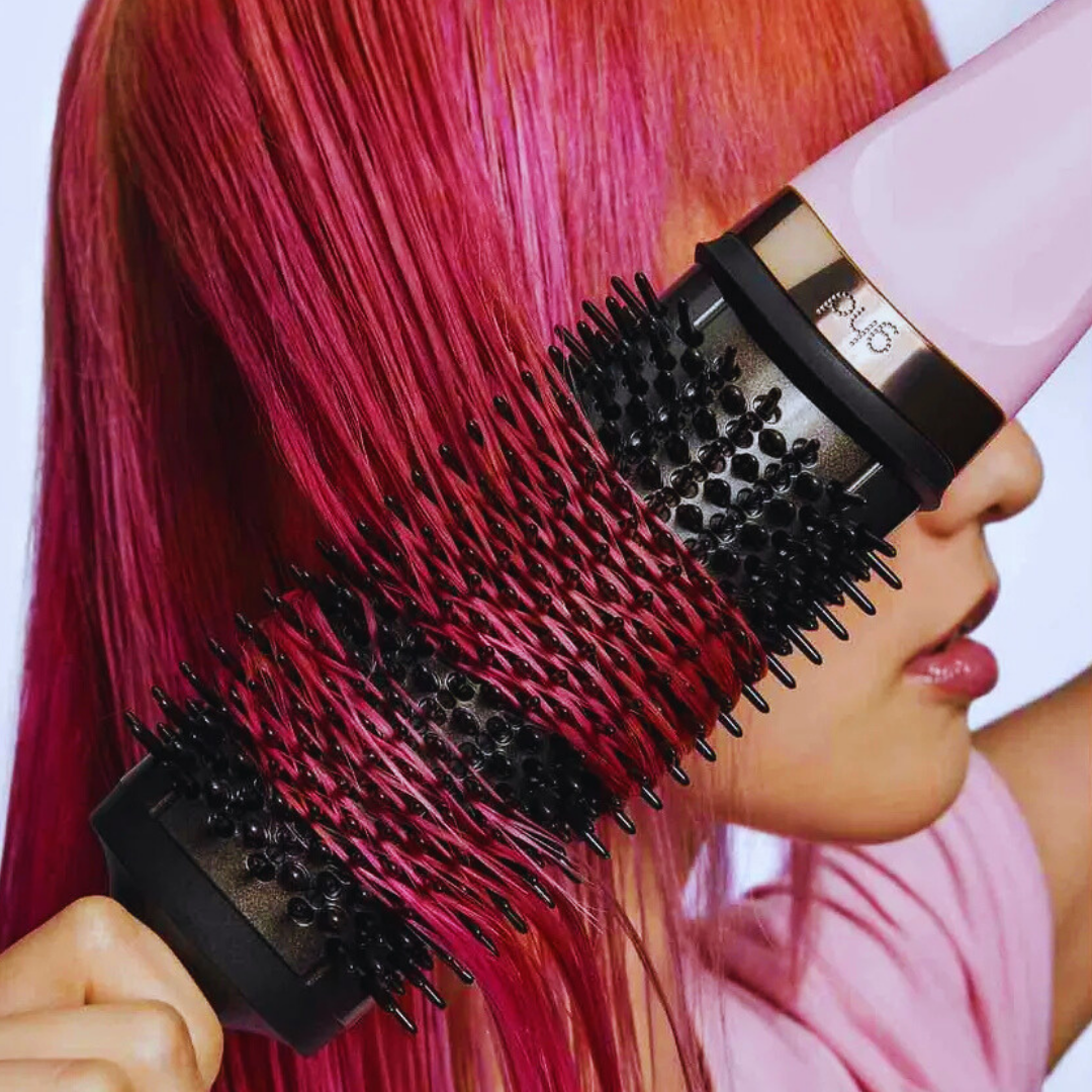 Eine Person benutzt einen rosa Haartrockner und eine Rundbürste auf pinkem Haar. Das Haar wird gestylt. Der Hintergrund ist weiß.