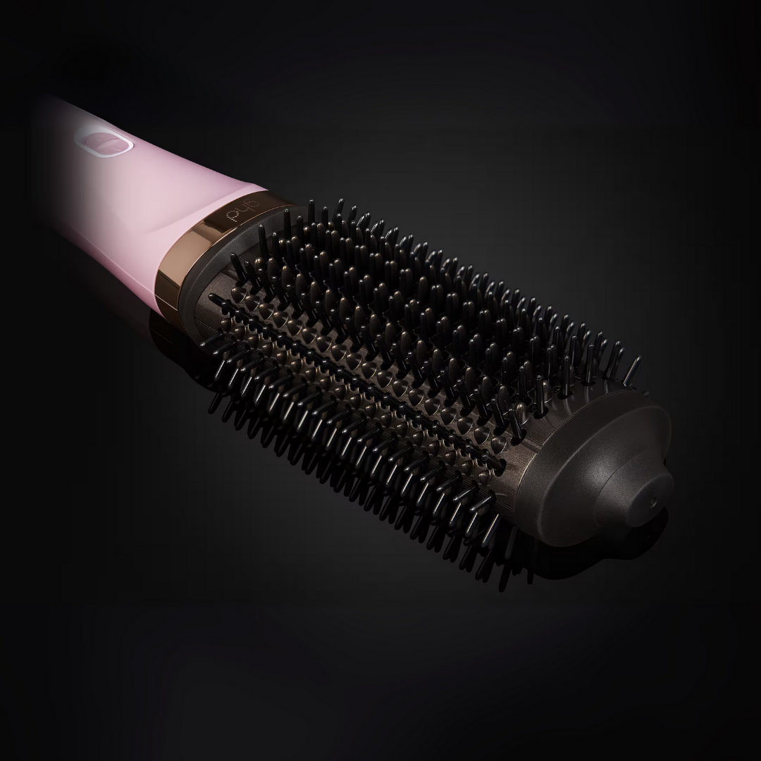 Eine rosa-goldene ghd Airbrush auf einer reflektierenden schwarzen Oberfläche. Die Bürste hat dunkle Borsten und ist nach rechts geneigt.
