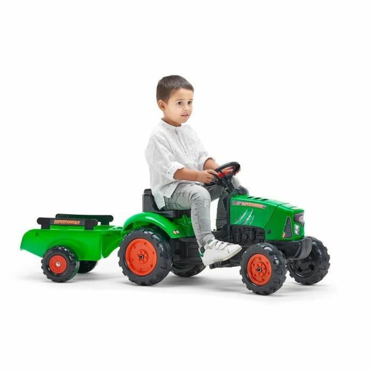 Ein Kind sitzt auf einem grünen Spielzeugtraktor mit Anhänger. Der Traktor hat orangefarbene Räder. Das Kind trägt ein weißes Hemd und graue Hosen.