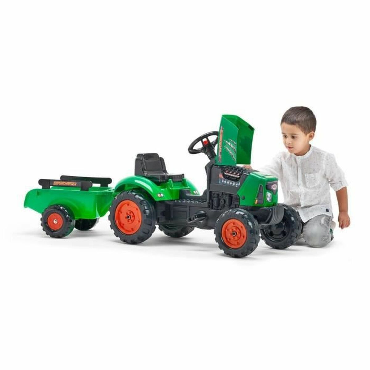 Ein Junge kniet neben einem grünen Spielzeugtraktor mit Anhänger. Der Traktor hat orangefarbene Räder. Der Junge trägt ein weißes Hemd.