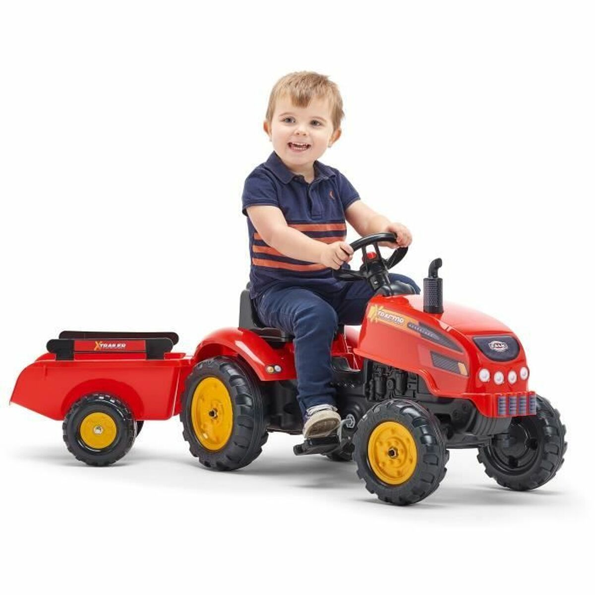 Ein lächelnder Junge fährt einen roten Spielzeugtraktor mit Anhänger. Der Traktor hat gelbe Räder, er trägt ein blaues Hemd und Jeans.