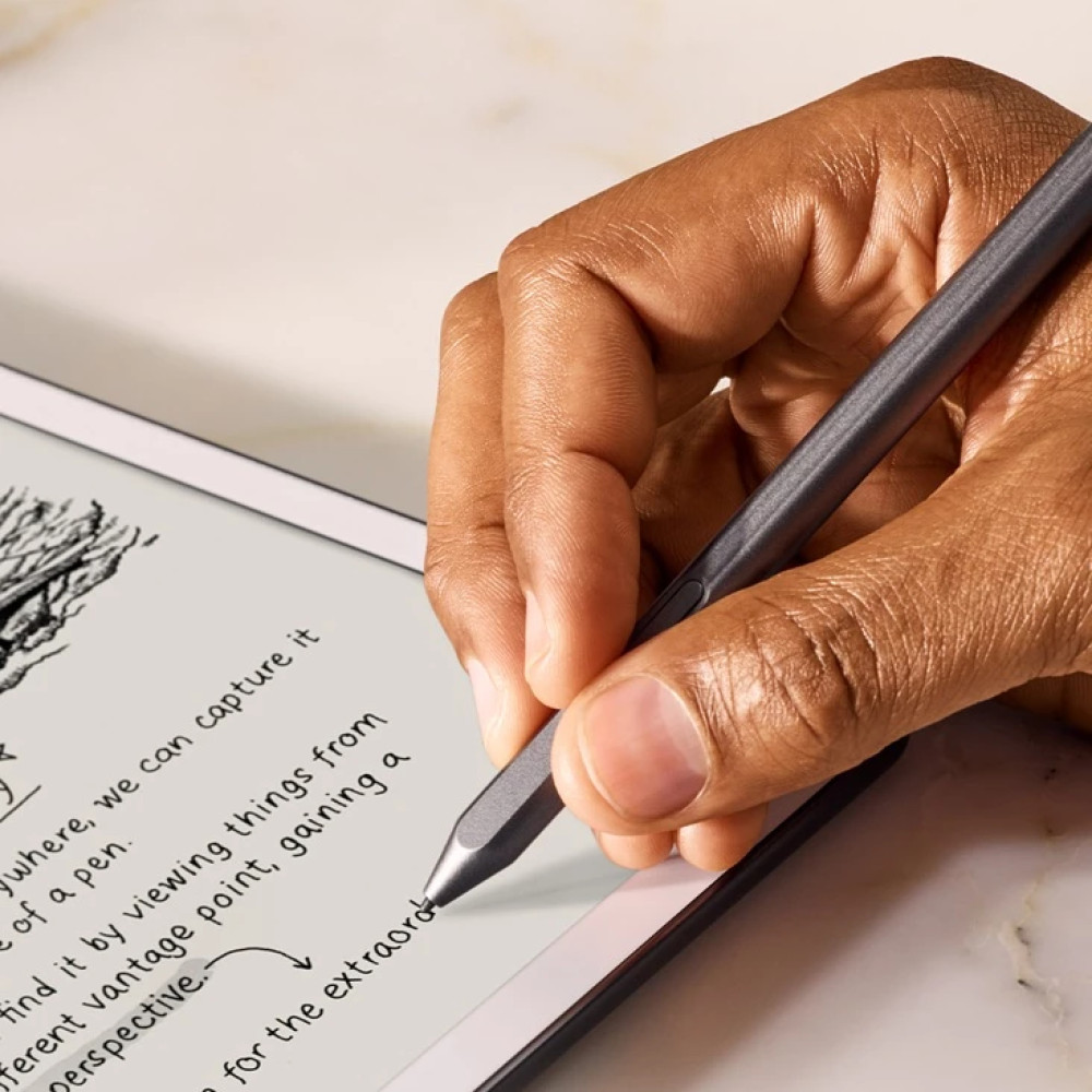 Ręka pisze na tablecie rysikiem. Tablet wyświetla tekst i mały szkic. Tło to marmurowa powierzchnia.