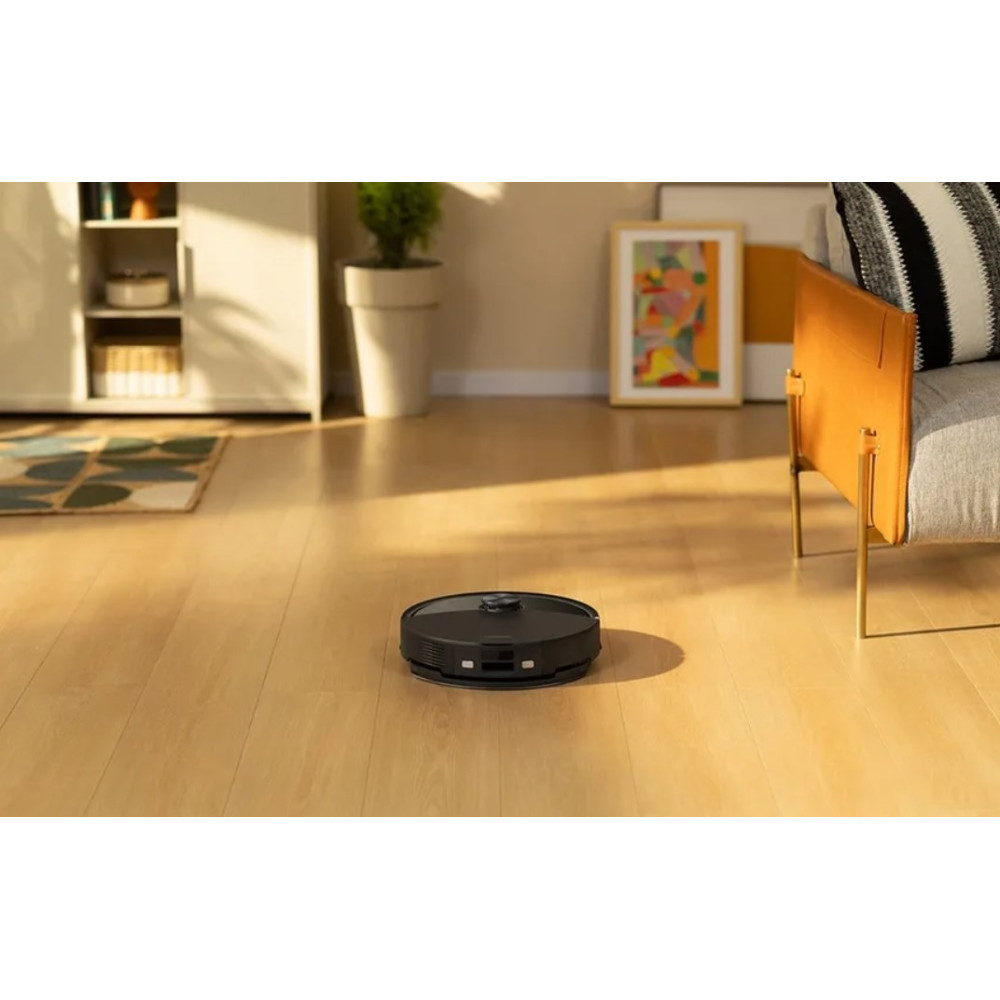 Un robot aspirapolvere nero su un pavimento in legno. Sullo sfondo un divano, un armadio, un dipinto e una pianta.