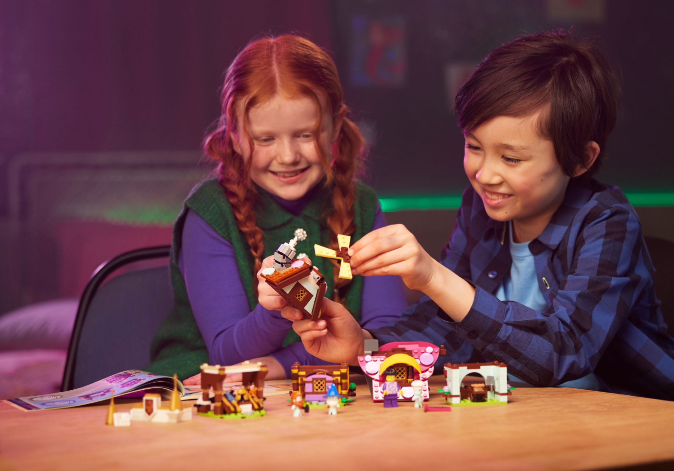 Dwoje dzieci składa Lego. Jedno trzyma element wiatraka. Inne zestawy Lego są na stole.