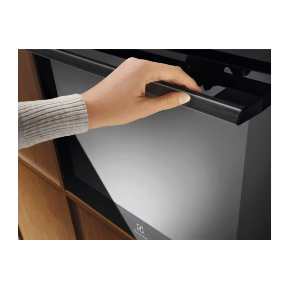 Ręka otwiera czarne drzwi piekarnika. Piekarnik jest wbudowany w drewnianą szafkę. Ręka ma na sobie szary sweter.