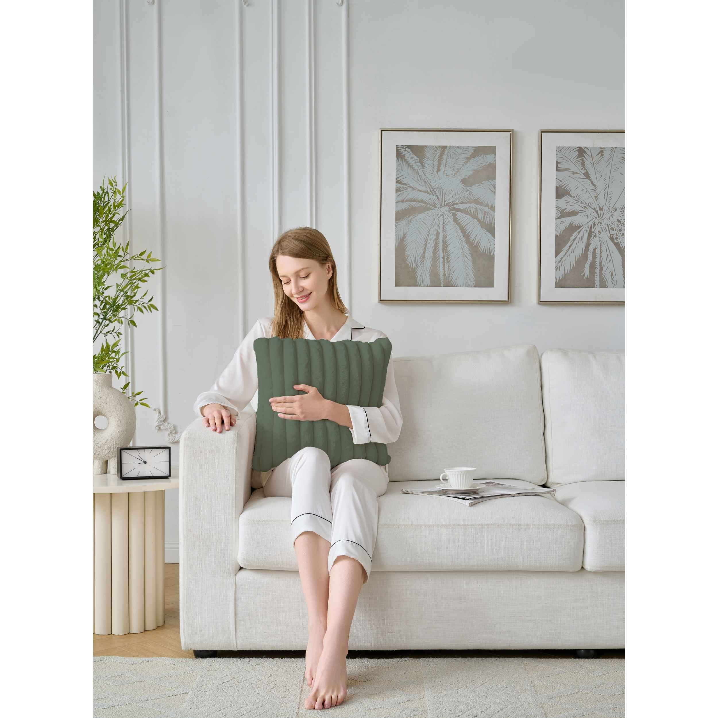 Eine Frau im Pyjama sitzt auf einem weißen Sofa und hält ein grünes Kissen. Palmenkunst hängt im Hintergrund.