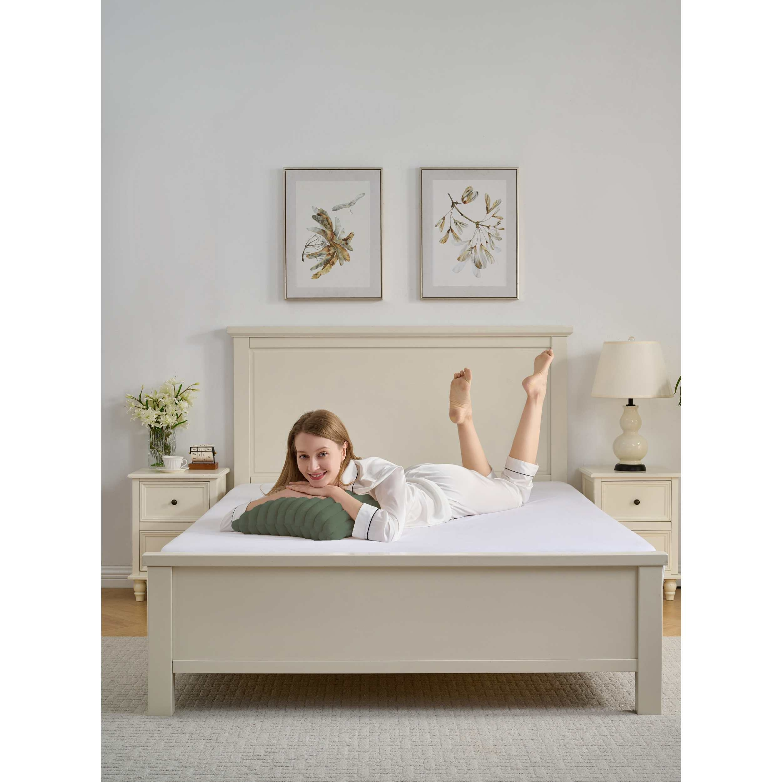 Eine Frau liegt lächelnd auf einem Bett. Sie trägt einen weißen Pyjama mit einem grünen Kissen. An der Wand hängen zwei gerahmte Kunstwerke.