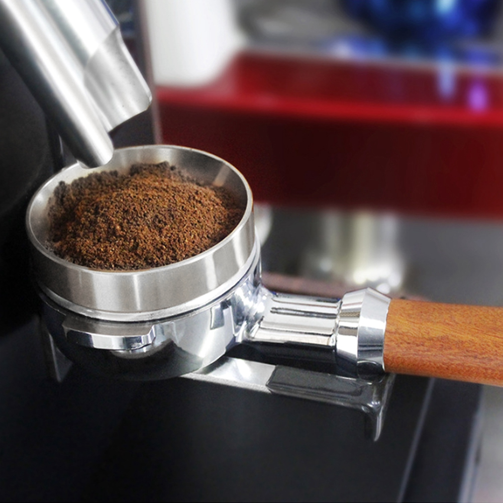 Nahaufnahme eines Siebträgers mit Kaffeepulver unter einer Mühle. Ein Holzgriff ist befestigt. Rotes Gerät im Hintergrund.