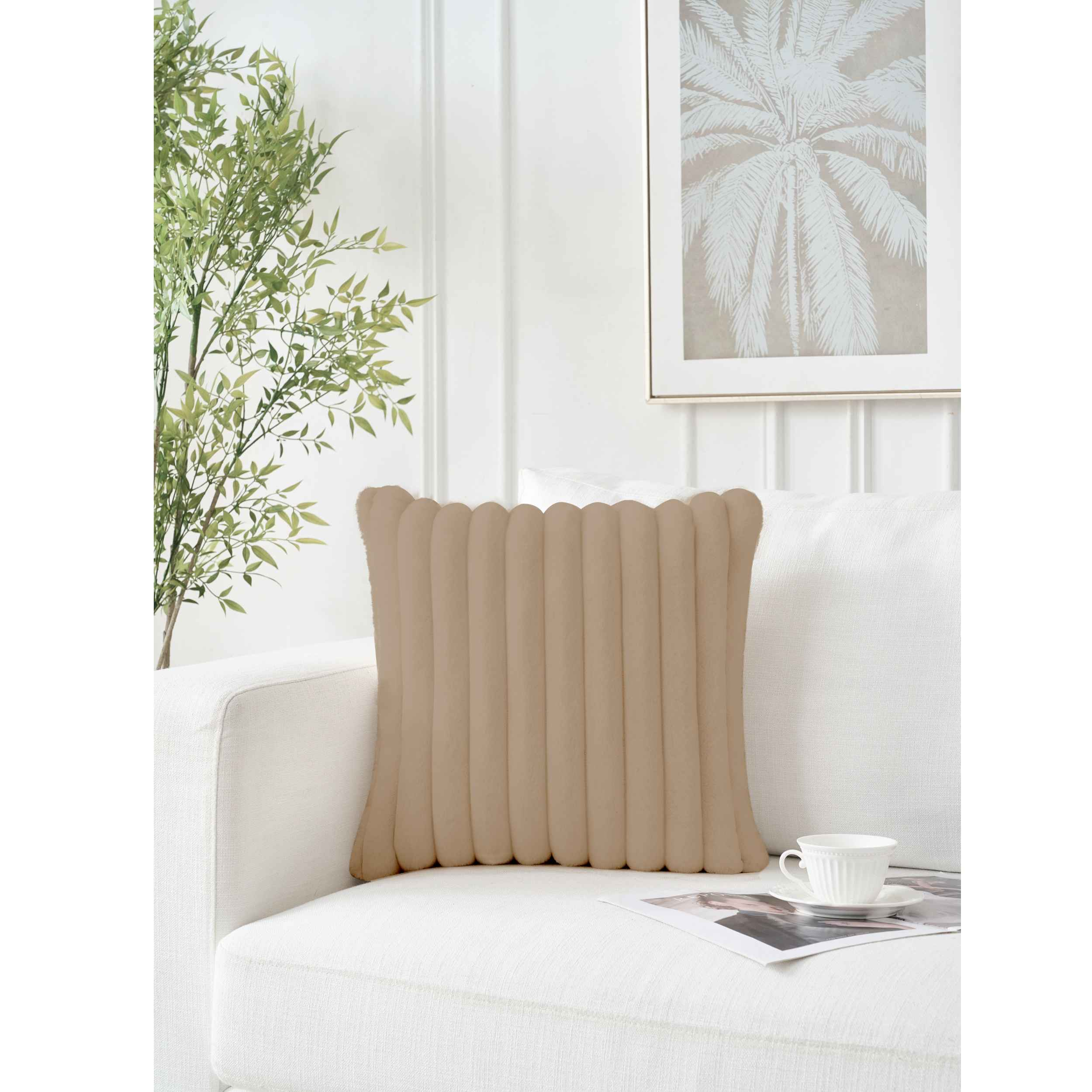 Ein beiges, geripptes Kissen liegt auf einer weißen Couch. Eine Teetasse und Kunst liegen auf der Couch. Palmenkunst und Pflanzen sind zu sehen.