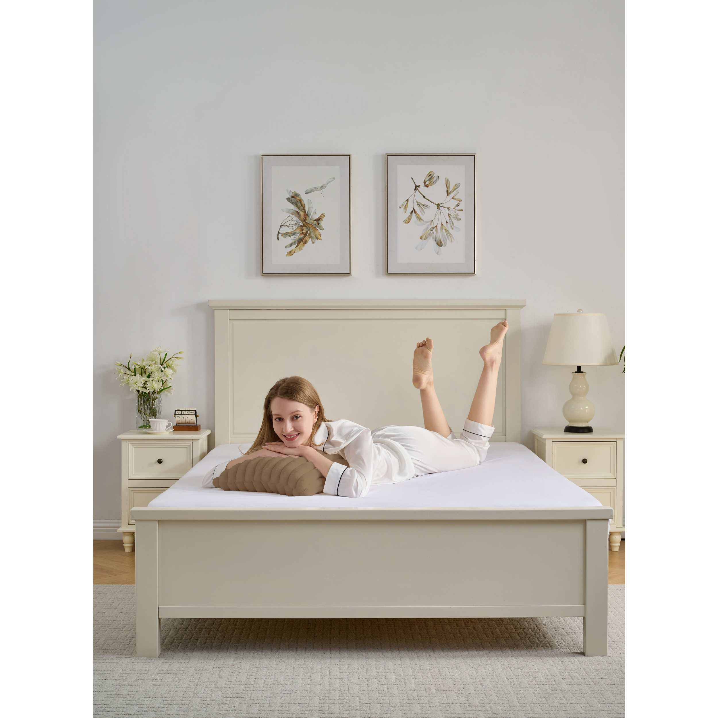 Eine Frau liegt lächelnd auf einem Bett. Sie trägt einen weißen Pyjama. An der Wand hängen Bilder und Nachttische.