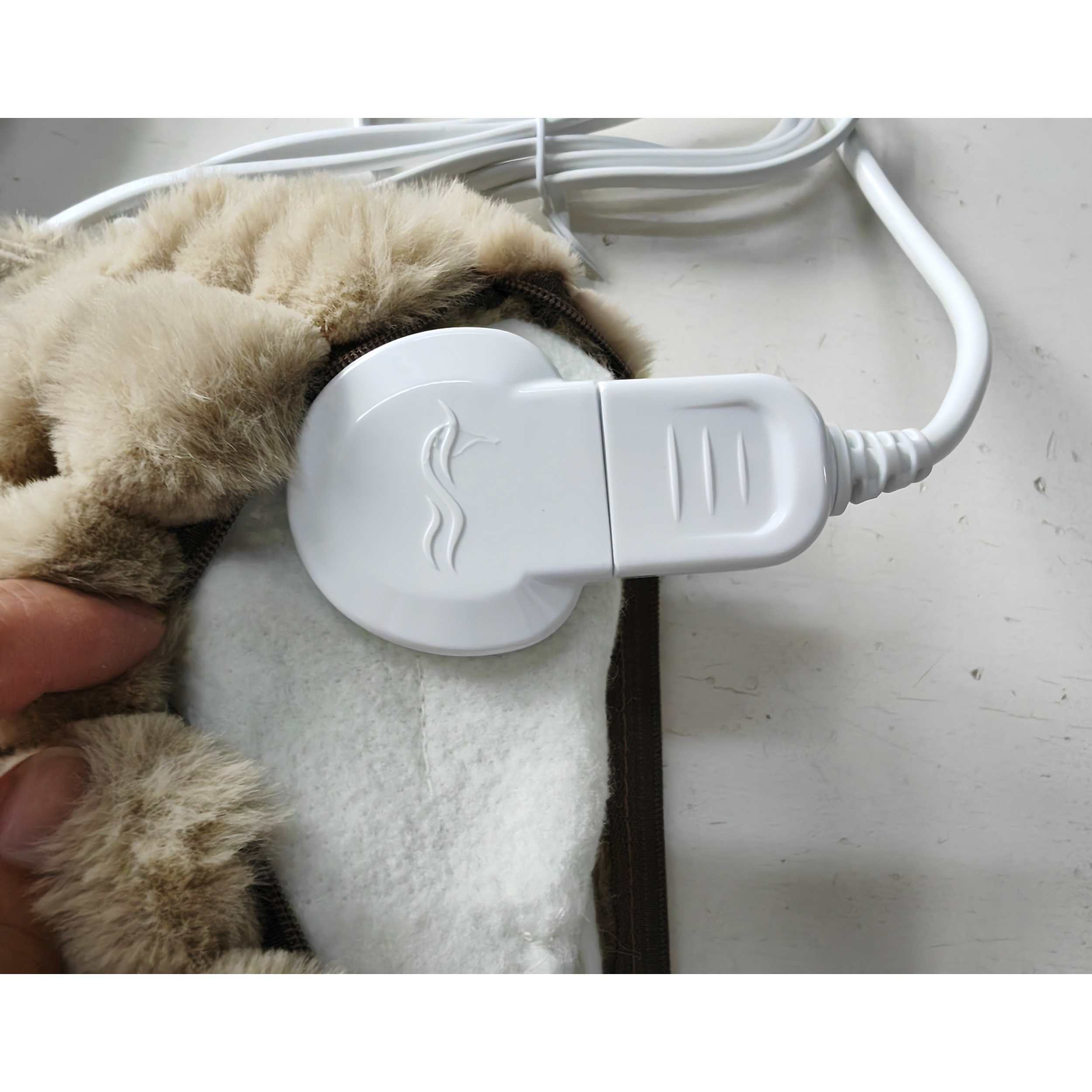Ein weißer Heizkissen-Controller ist mit einer beigefarbenen, fellartigen Abdeckung verbunden. Eine Hand berührt das Kissen.
