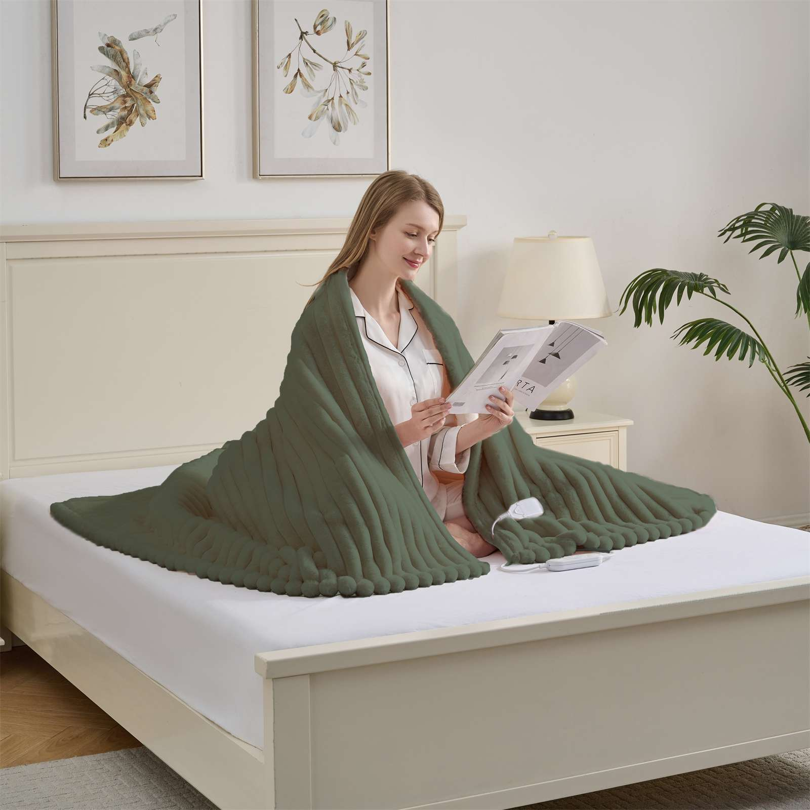 Eine Frau sitzt auf einem Bett, in eine grüne Decke gehüllt, und liest eine Zeitschrift. Eine Lampe, eine Pflanze und Kunstwerke sind im Hintergrund.