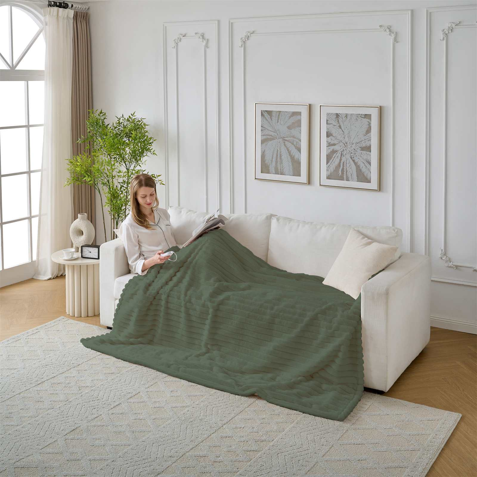 Frau auf weißem Sofa mit grüner Decke, liest. Zwei gerahmte Bilder und ein Fenster sind sichtbar.