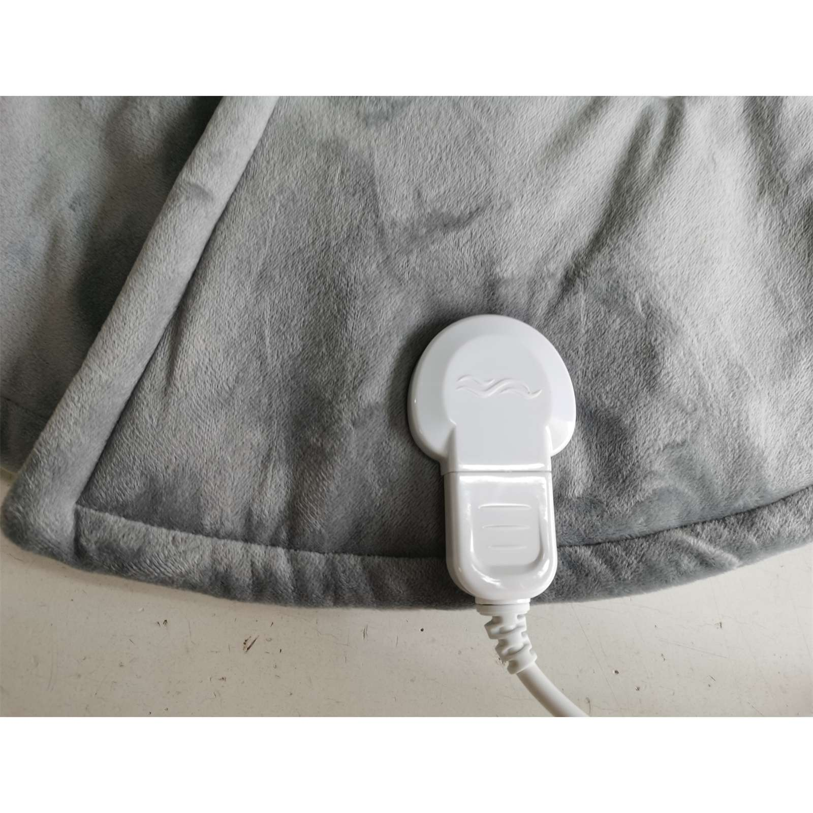 Eine graue Heizdecke mit weißem Stecker. Die Decke liegt auf einer weißen Oberfläche.