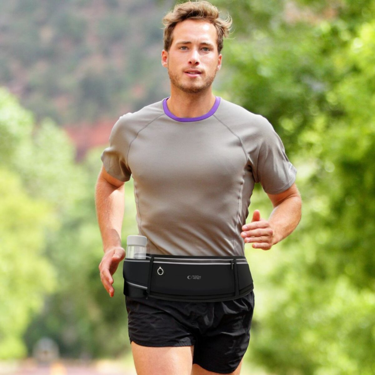Ein Mann läuft im Freien, trägt ein graues Hemd, schwarze Shorts und einen Laufgürtel mit einer Wasserflasche. Grünes Laub im Hintergrund.