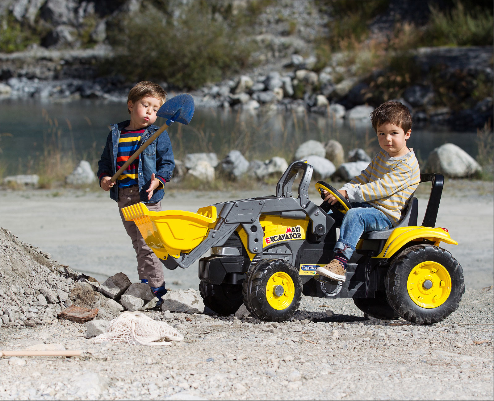 Dos niños juegan con un juguete excavadora amarilla. Uno sostiene una pala azul, el otro está en el asiento del conductor. Rocas y agua al fondo.