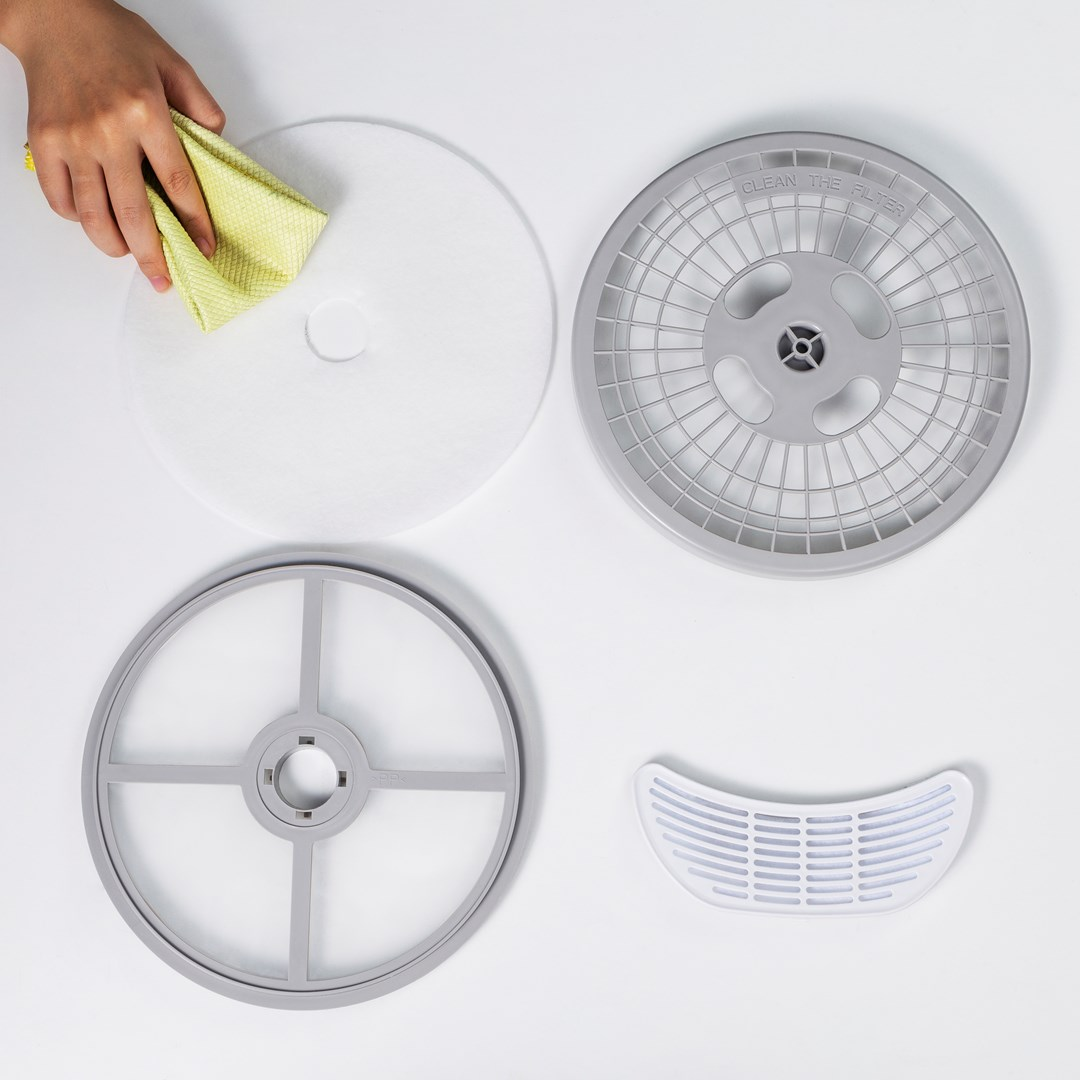 Ręka czyści biały filtr żółtą szmatką. Elementy oczyszczacza powietrza: szara rama, szara kratka i biały filtr.