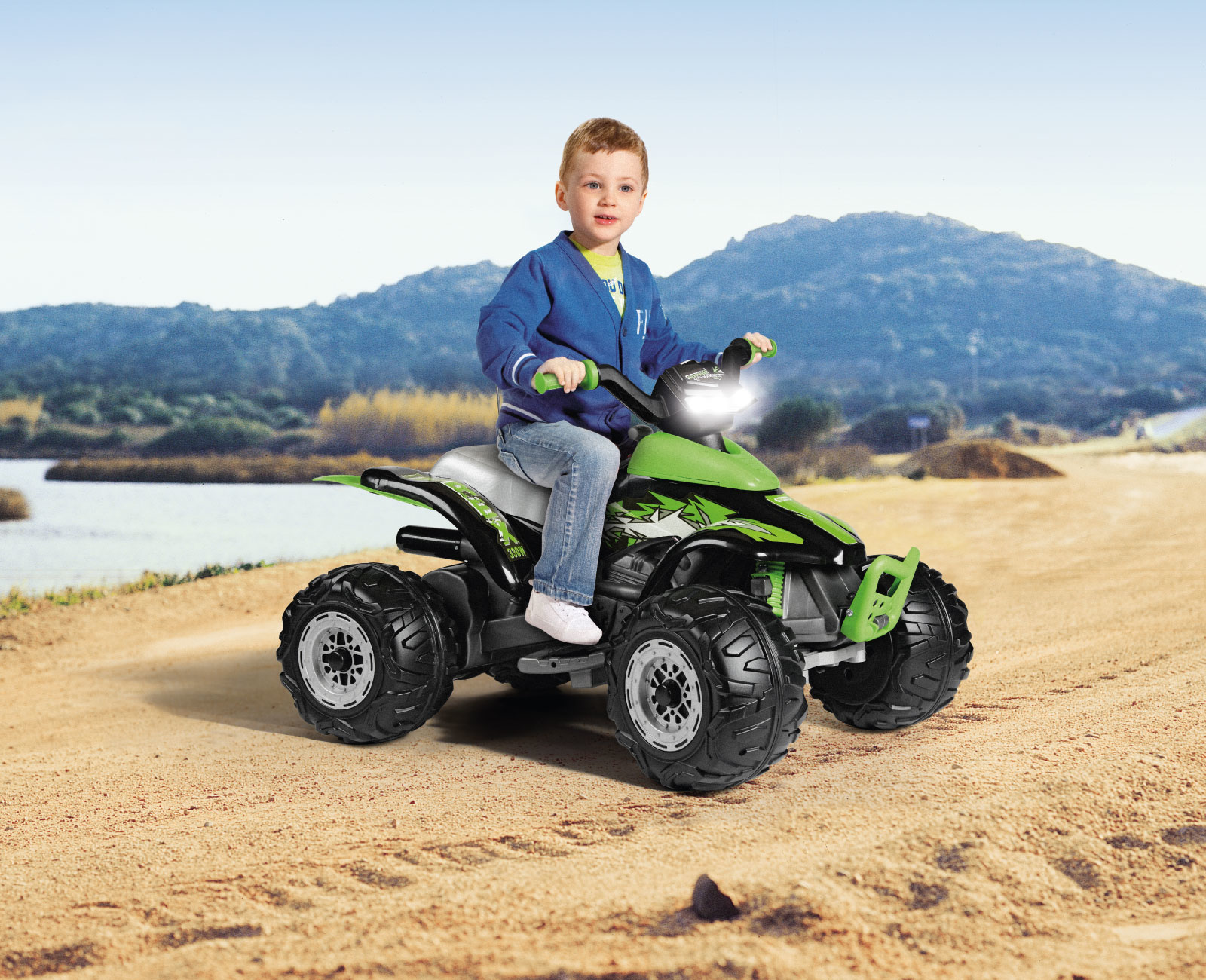Un bambino piccolo è seduto su un quad verde e nero su una superficie sabbiosa, con montagne sullo sfondo.