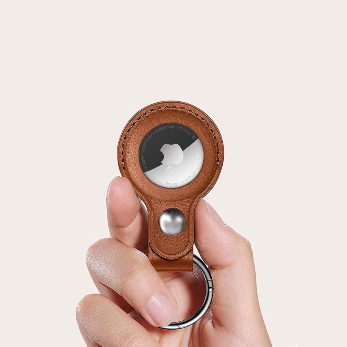 Eine Hand hält einen braunen Leder-AirTag-Halter. Der AirTag ist weiß mit einem schwarzen Apple-Logo. Ein silberner Schlüsselring ist befestigt.