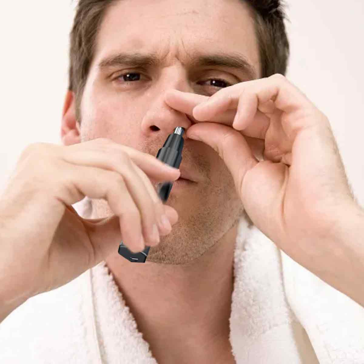 Ein Mann trimmt Nasenhaare mit einem schwarzen Trimmer. Er trägt einen weißen Bademantel. Nahaufnahme von Gesicht, Händen und Trimmer.