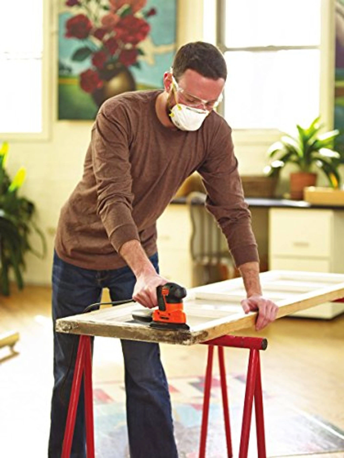 Mężczyzna szlifuje drewnianą ościeżnicę, nosząc maskę i okulary ochronne. Używa pomarańczowej szlifierki. Widoczny jest obraz i roślina.