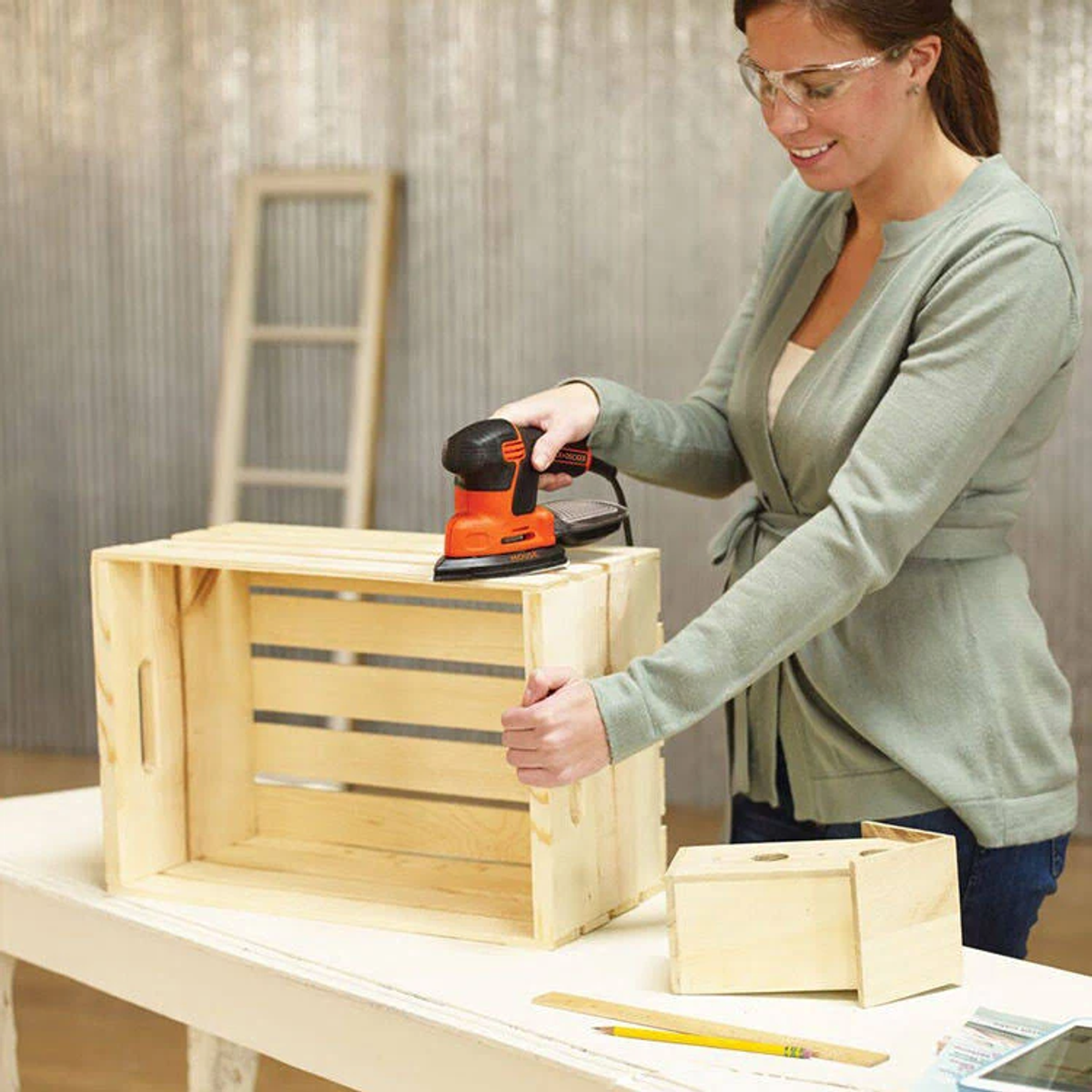 Kobieta szlifuje drewnianą skrzynię szlifierką elektryczną. Na stole znajduje się małe drewniane pudełko, linijka i ołówek.