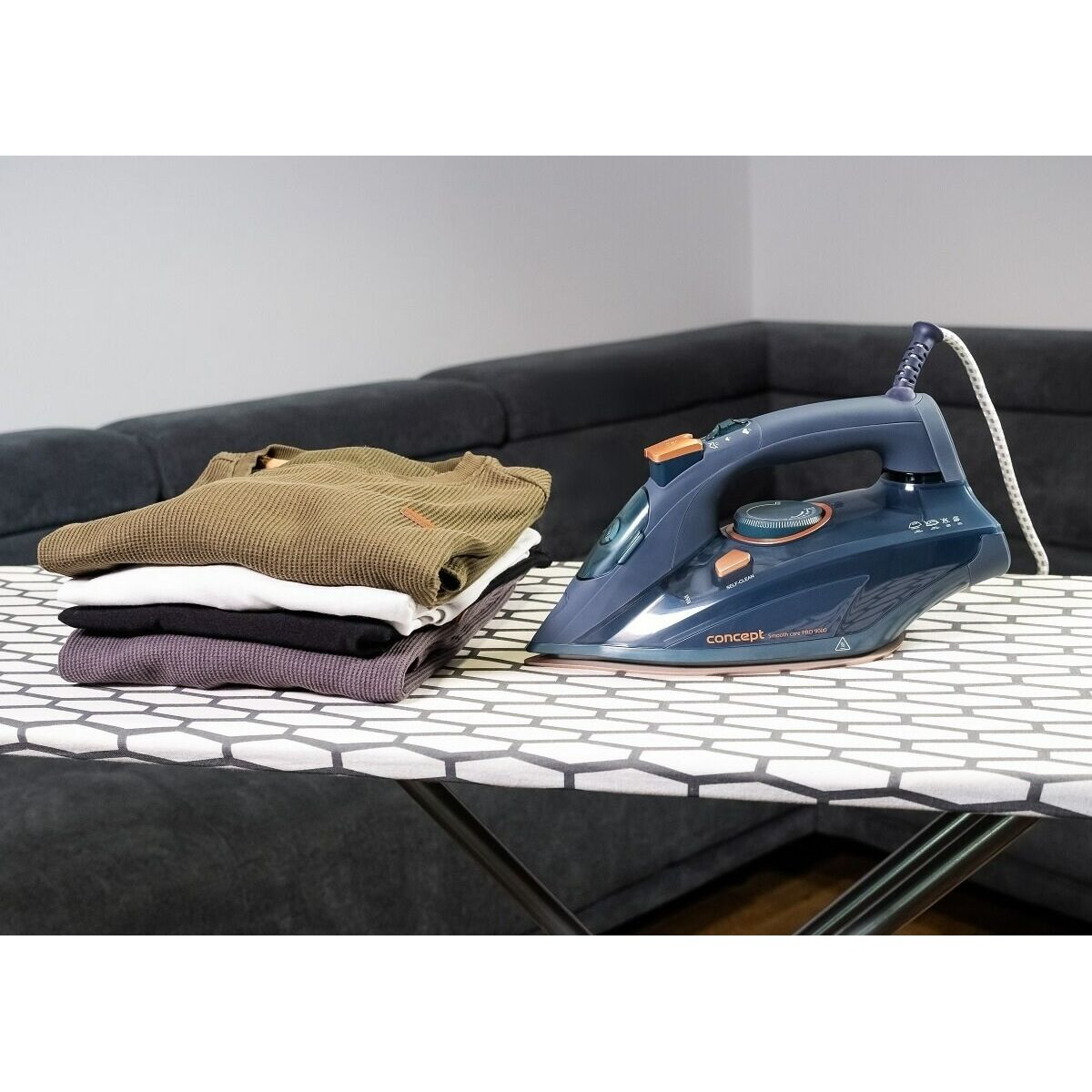 Deska do prasowania z niebieskim żelazkiem, stos złożonych koszul i szara sofa w tle.