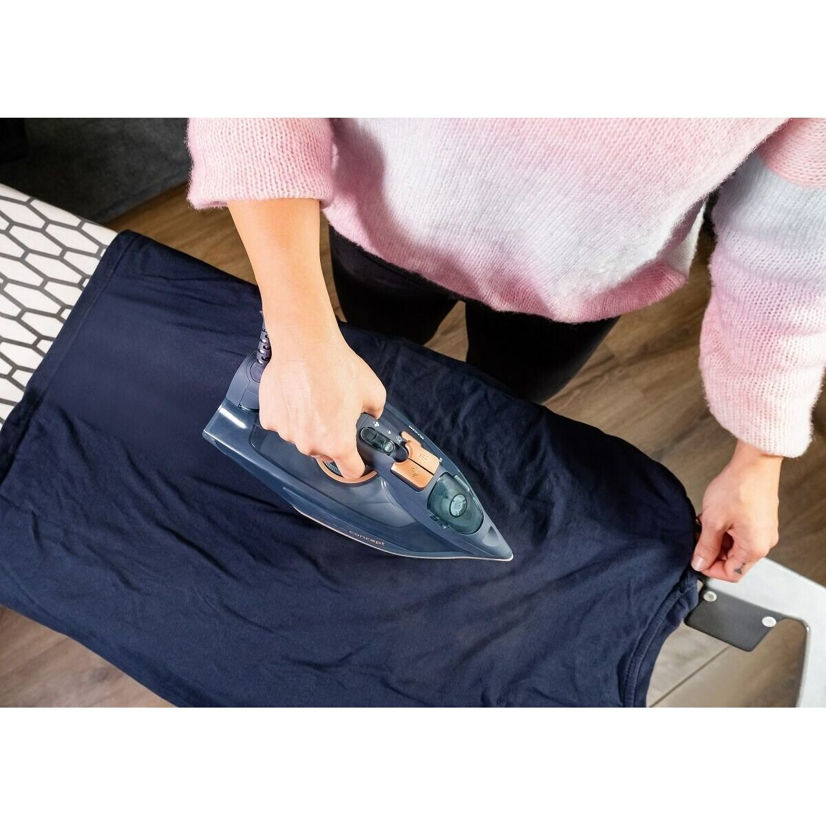 Osoba prasuje granatową koszulkę niebieskim żelazkiem. Żelazko znajduje się na desce do prasowania. Osoba ma na sobie różowy sweter.