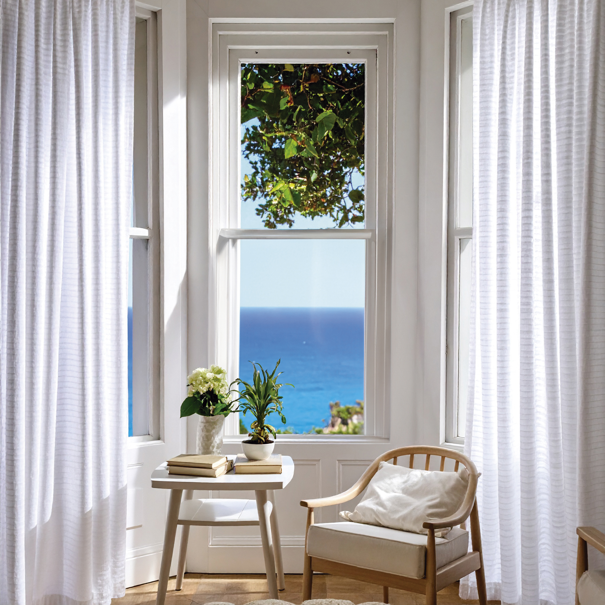 Heller Raum mit Fenster mit Meerblick. Weiße Vorhänge, Tisch mit Pflanzen, Stuhl mit Kissen.