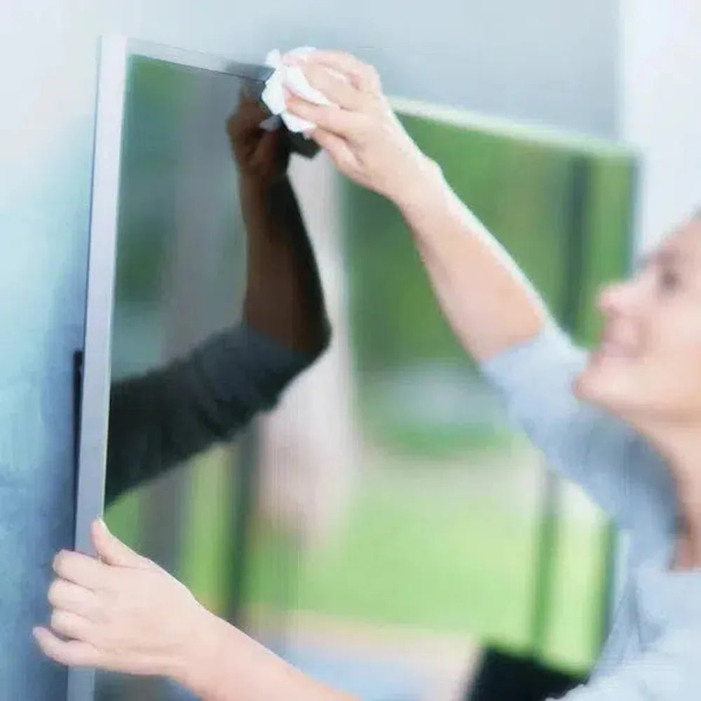 A person cleaning a large TV screen with a white cloth. The screen reflects a green outdoor scene.