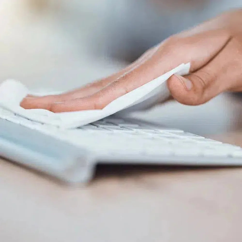 A hand wipes a white keyboard with a white wipe. The keyboard is on a light wooden desk. Focus is on the cleaning.