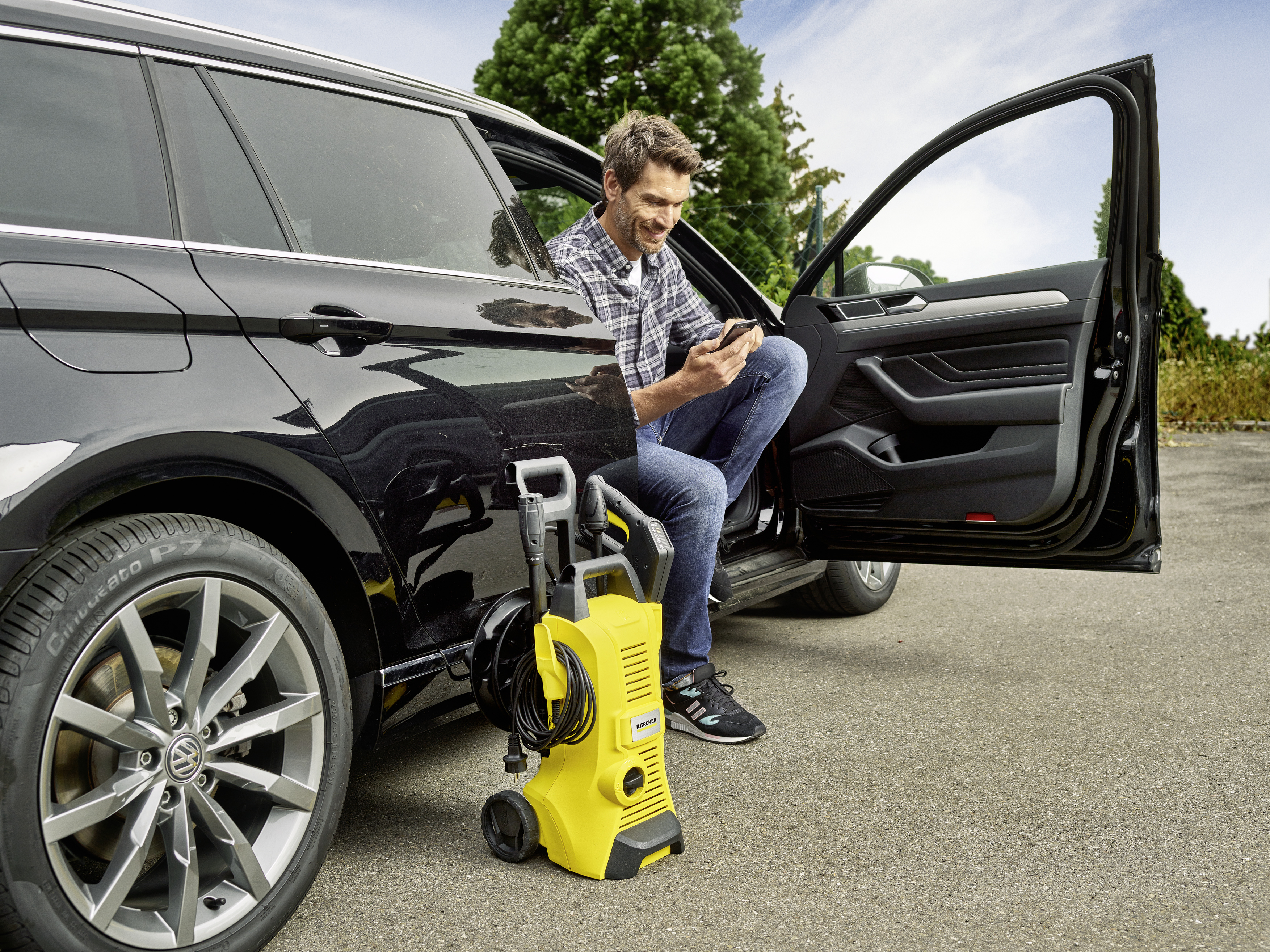 Mężczyzna w samochodzie z telefonem, żółta myjka ciśnieniowa na zewnątrz. Czarny samochód na asfalcie. Zielone drzewa i niebieskie niebo.