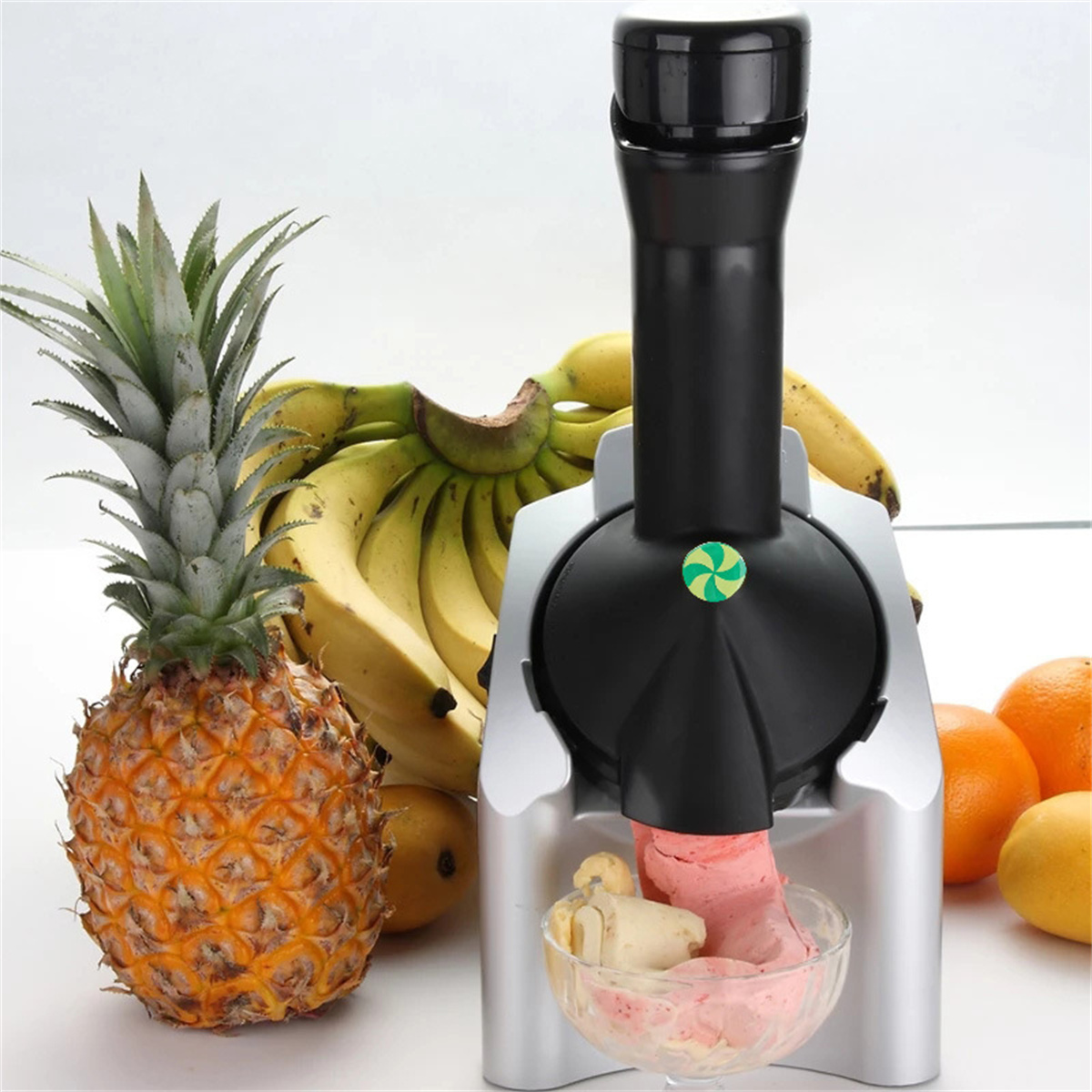 Eine Fruchteismaschine mit Ananas, Bananen und Orangen. Die Maschine gibt rosa und weißes Eis in eine Glasschale ab.