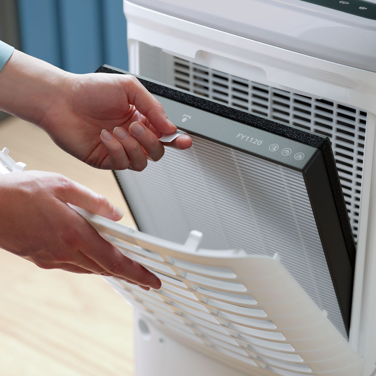 Ręce wyjmują filtr z białego oczyszczacza powietrza. Filtr jest czarny i szary, z białą osłoną ochronną.