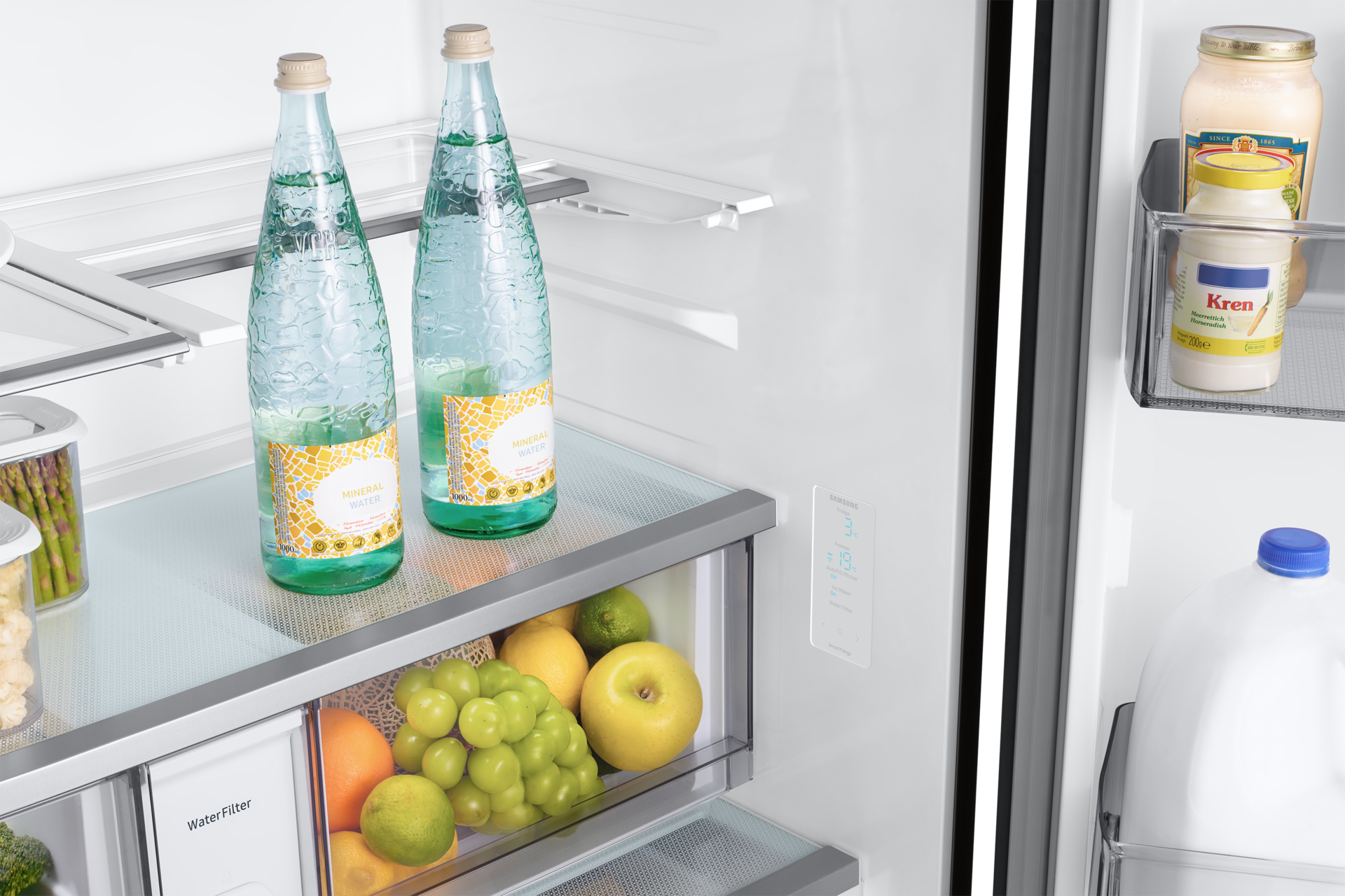 In een koelkast: mineraalwaterflessen, fruit (druiven, citroen, appel), kruiden en een melkkan. De koelkast is schoon en goed georganiseerd.