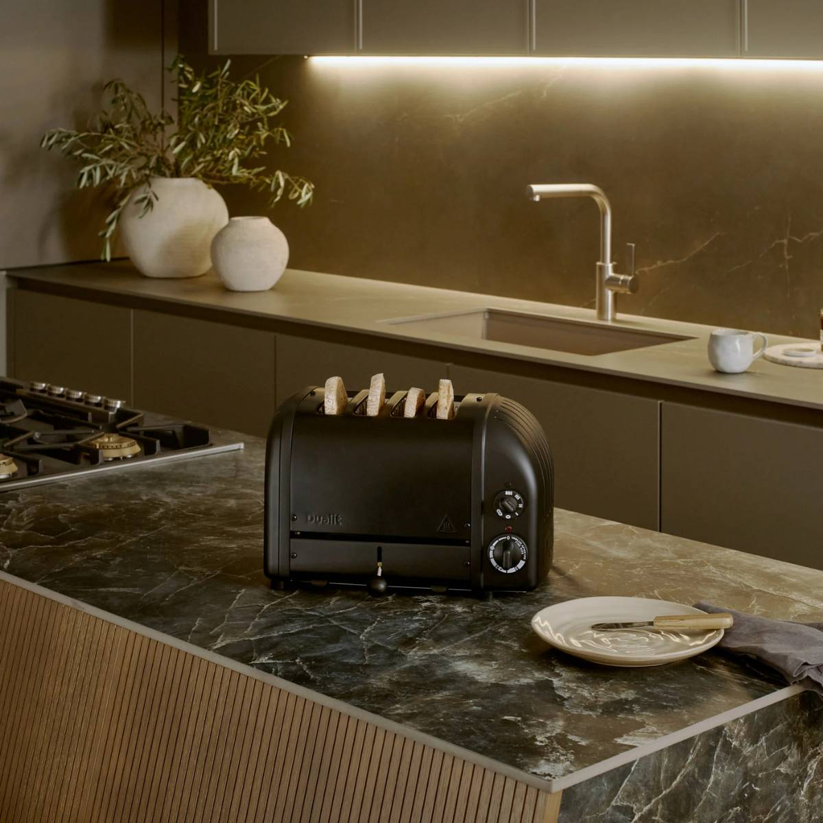 Ein schwarzer Toaster mit Brotscheiben steht auf einer Marmorarbeitsplatte. Ein Teller mit Messer und ein Waschbecken sind ebenfalls zu sehen.
