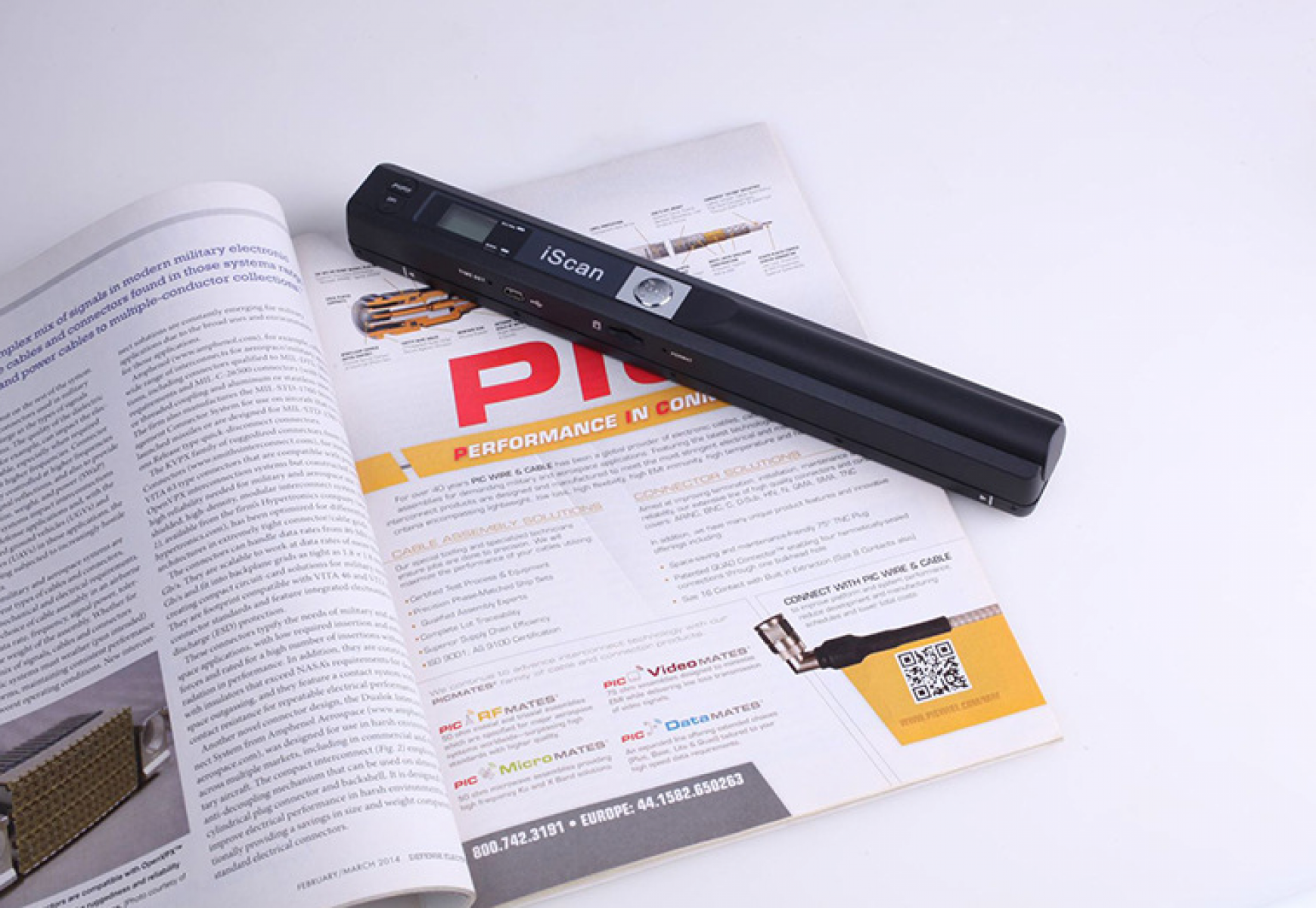 Ein schwarzer iScan-Scanner auf einem offenen Magazin. Das Magazin zeigt Text und Grafiken mit roten Akzenten. Weiße Oberfläche.