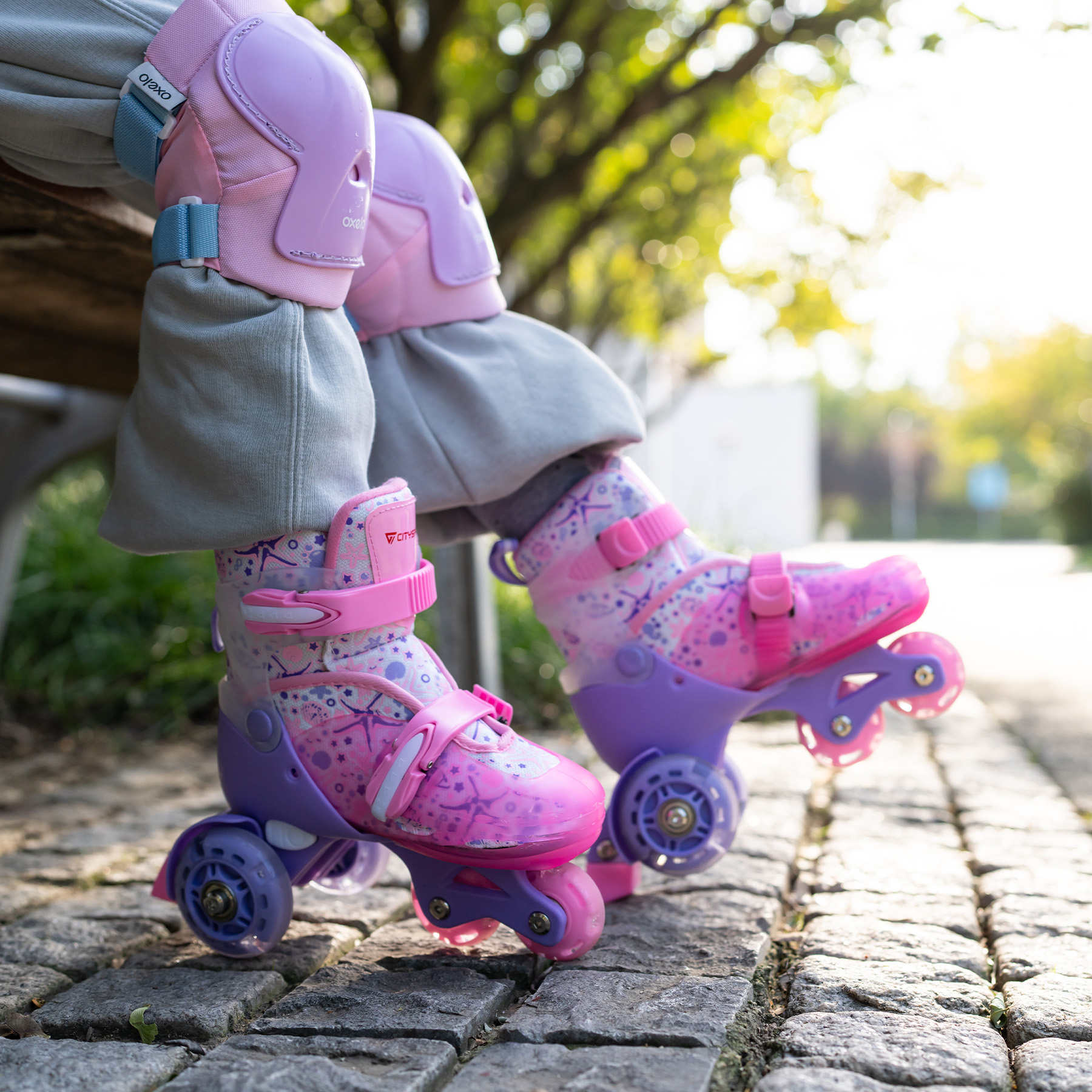 Die Beine eines Kindes mit pinkfarbenen und lila Rollschuhen. Rosa Knieschützer sind sichtbar. Szene auf einem Kopfsteinpflasterweg.