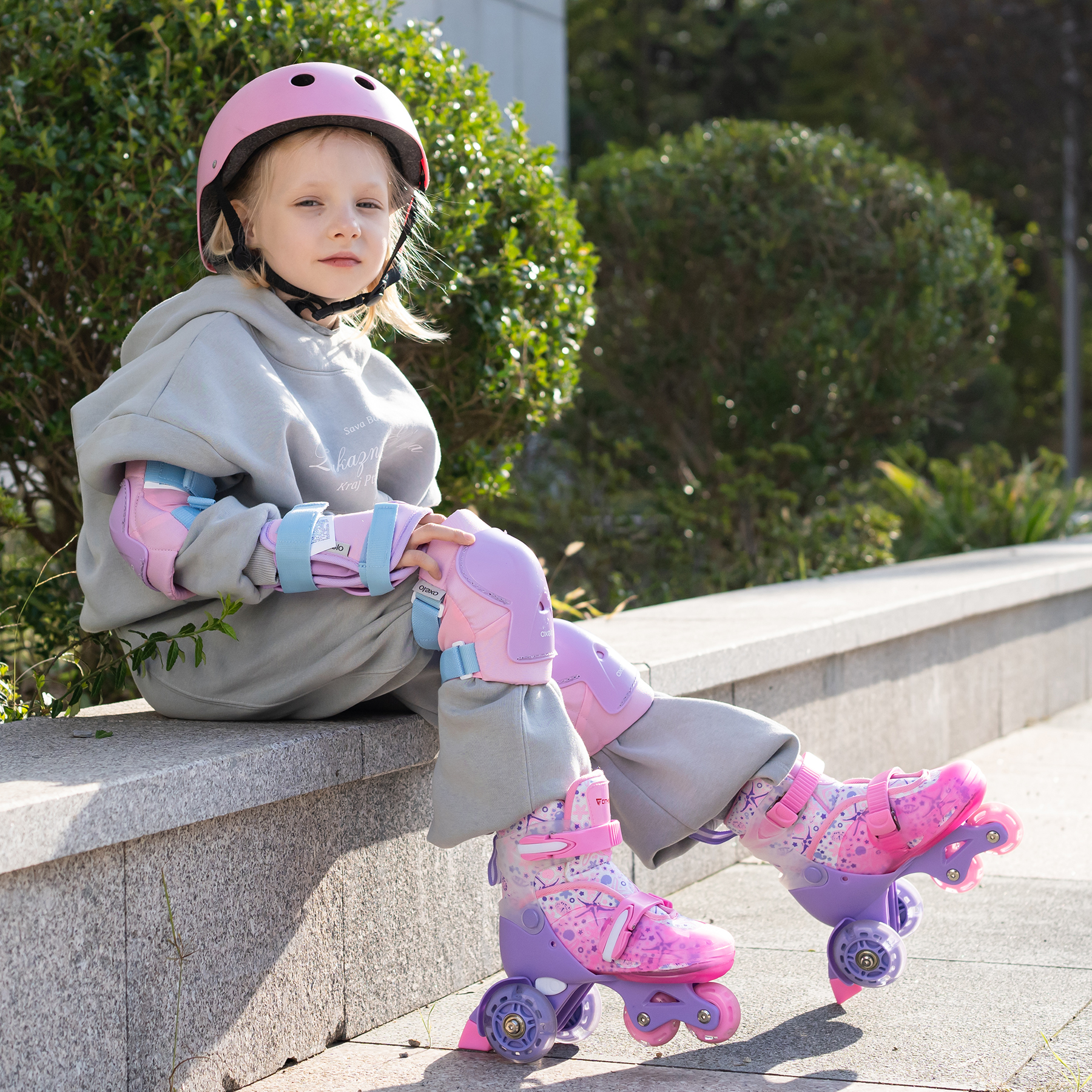 Ein Mädchen mit rosa Helm und Rollschuhen sitzt auf einer Mauer und trägt Schutzkleidung. Im Hintergrund grüne Büsche.