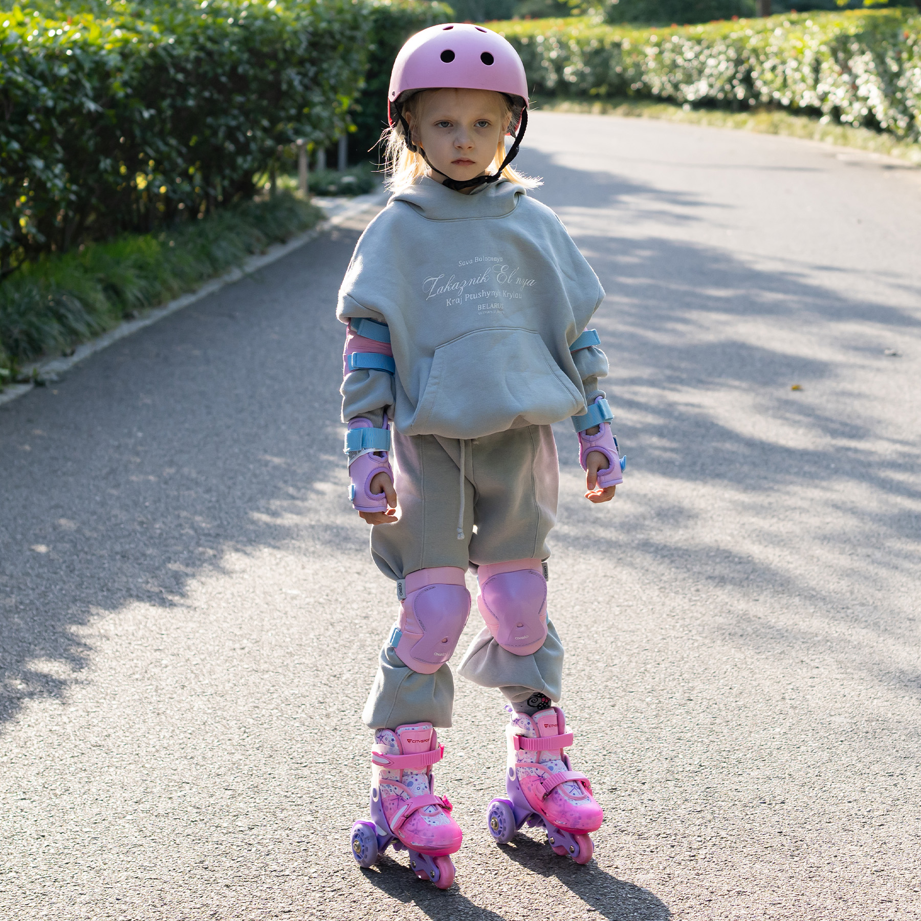 Ein Mädchen mit Schutzkleidung und Rollschuhen steht auf einem Weg. Sie trägt einen rosa Helm und einen grauen Trainingsanzug.
