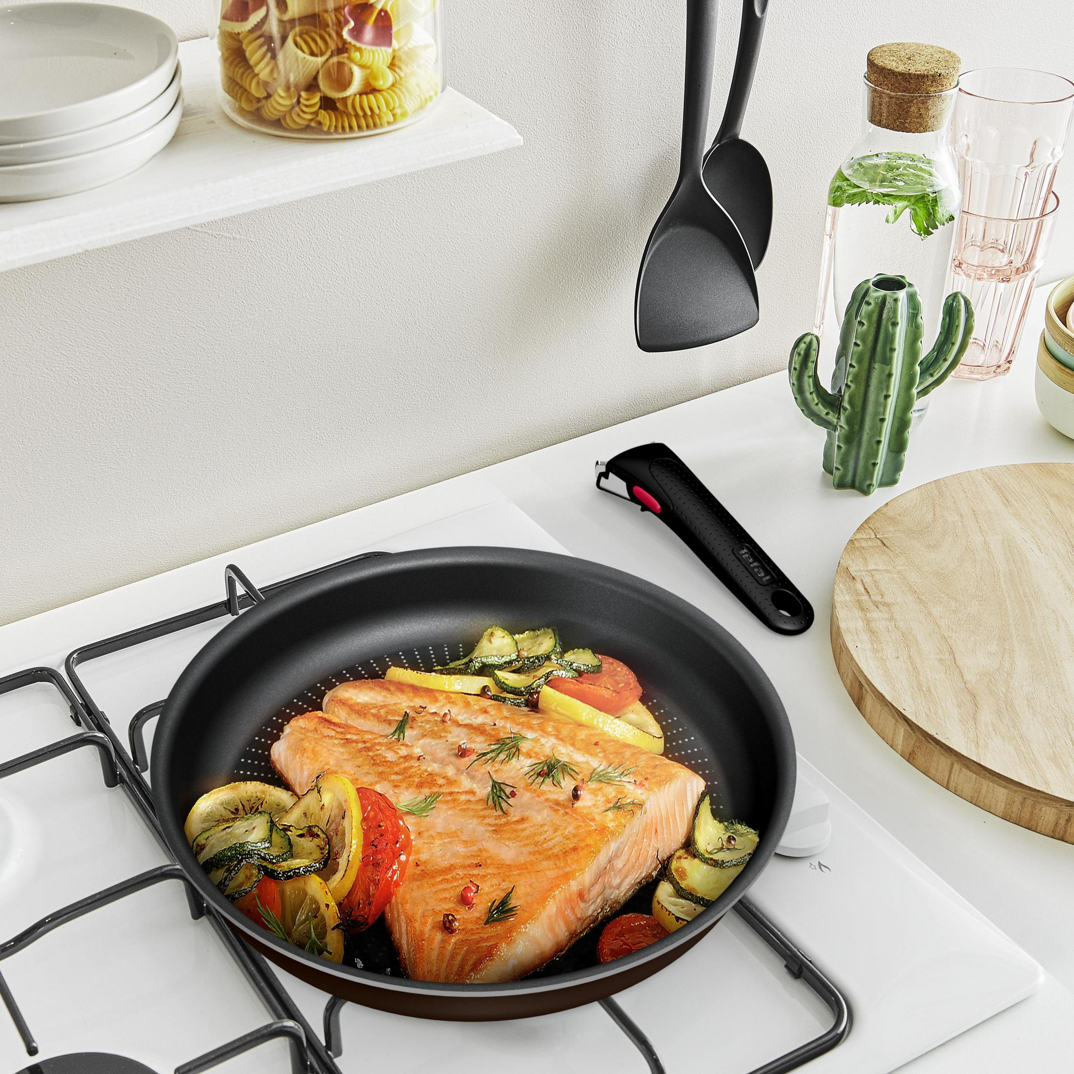 Une poêle avec du saumon et des légumes est sur une cuisinière. Il y a des ustensiles de cuisine.