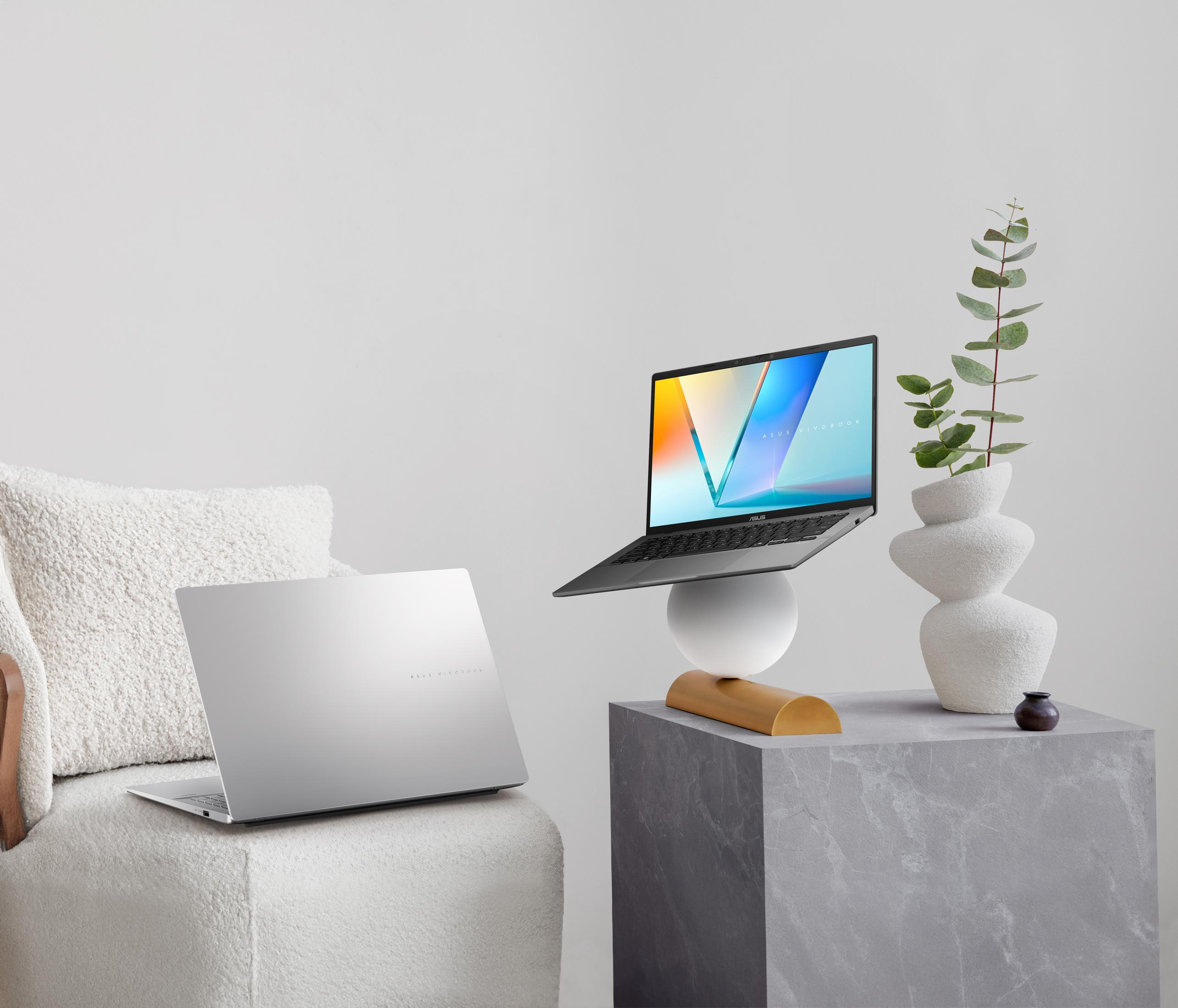 Dwa srebrne laptopy, jeden na białej kuli i drewnie, drugi na białym fotelu. Wazon z rośliną na kostce.