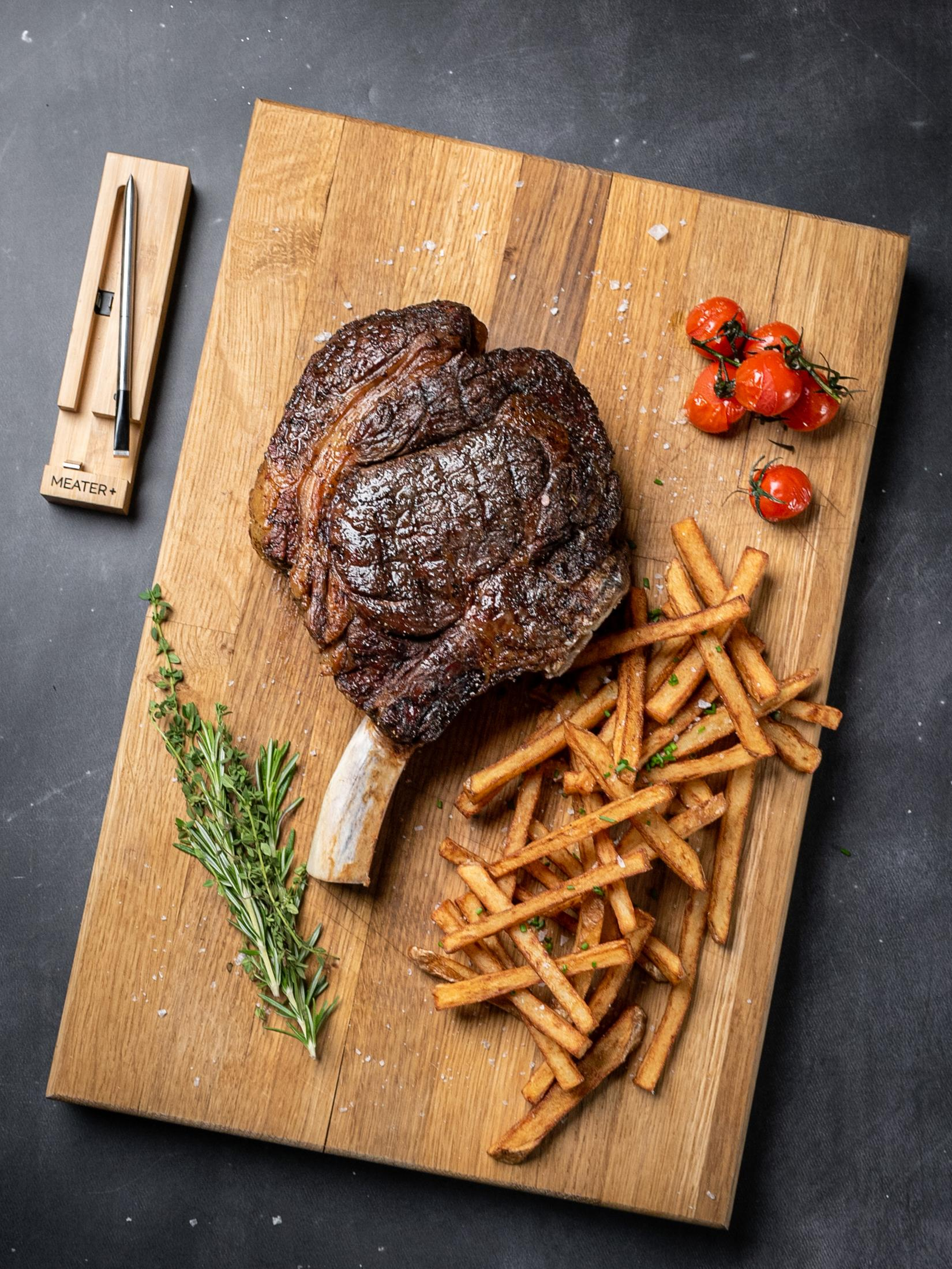Gegrilltes Steak mit Knochen, Pommes, Tomaten und Kräutern auf einem Holzbrett. Ein Fleischthermometer liegt daneben.