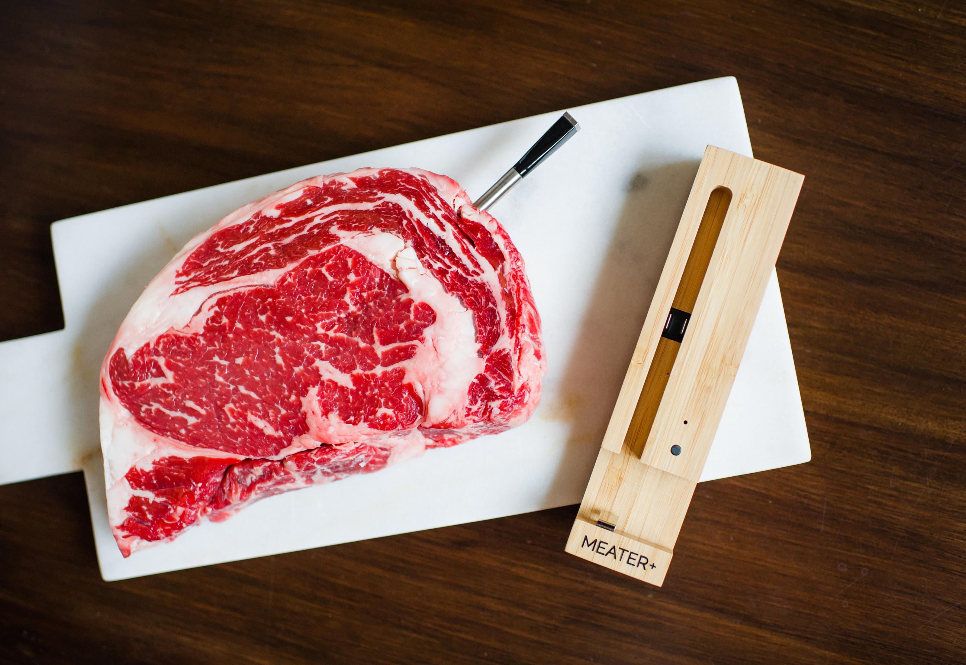 Ein rohes Ribeye-Steak mit Fleischthermometer auf einem weißen Brett mit einem hölzernen Thermometerhalter.