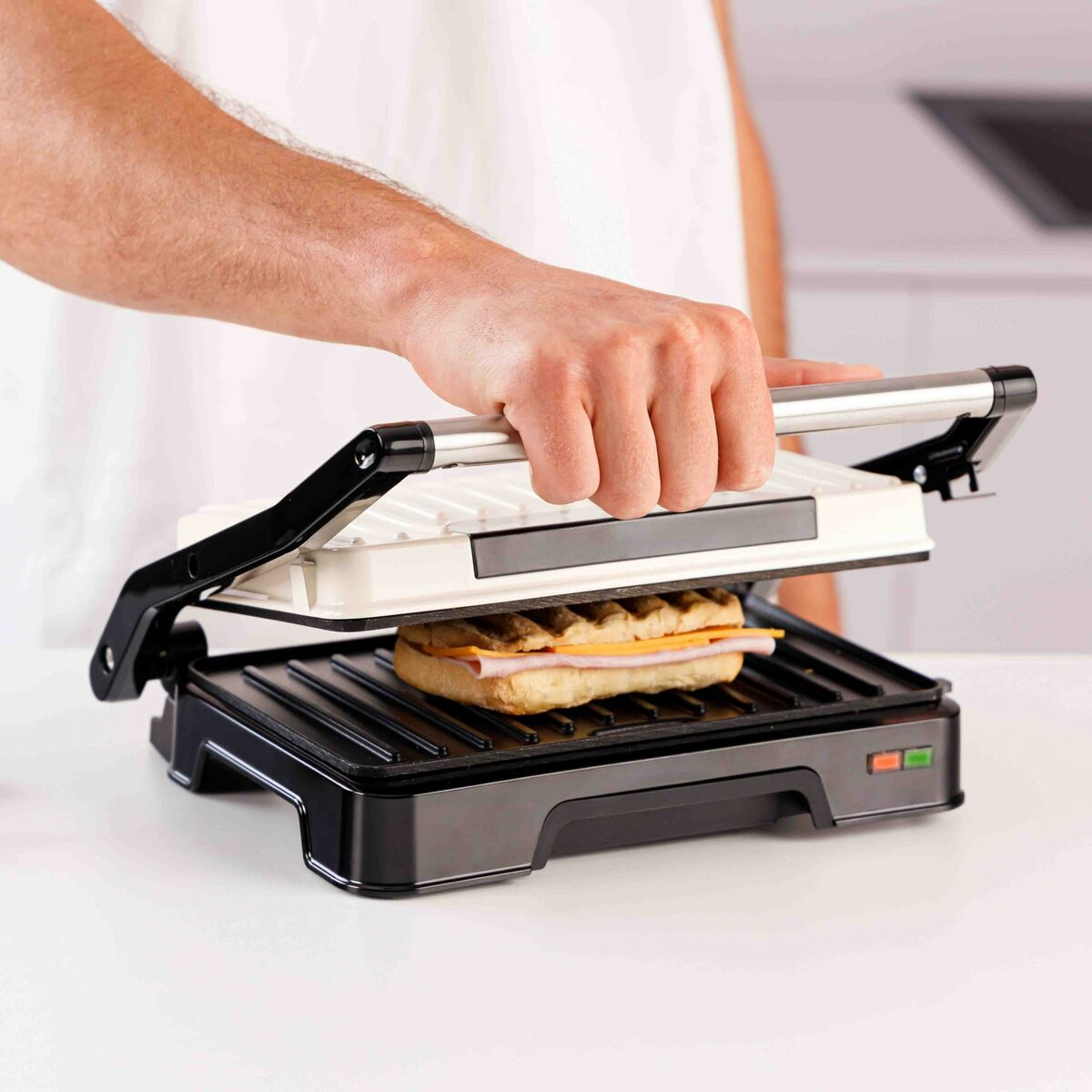 Una persona usa una piastra per panini per cucinare un panino con prosciutto e formaggio. La piastra è bianca e nera.
