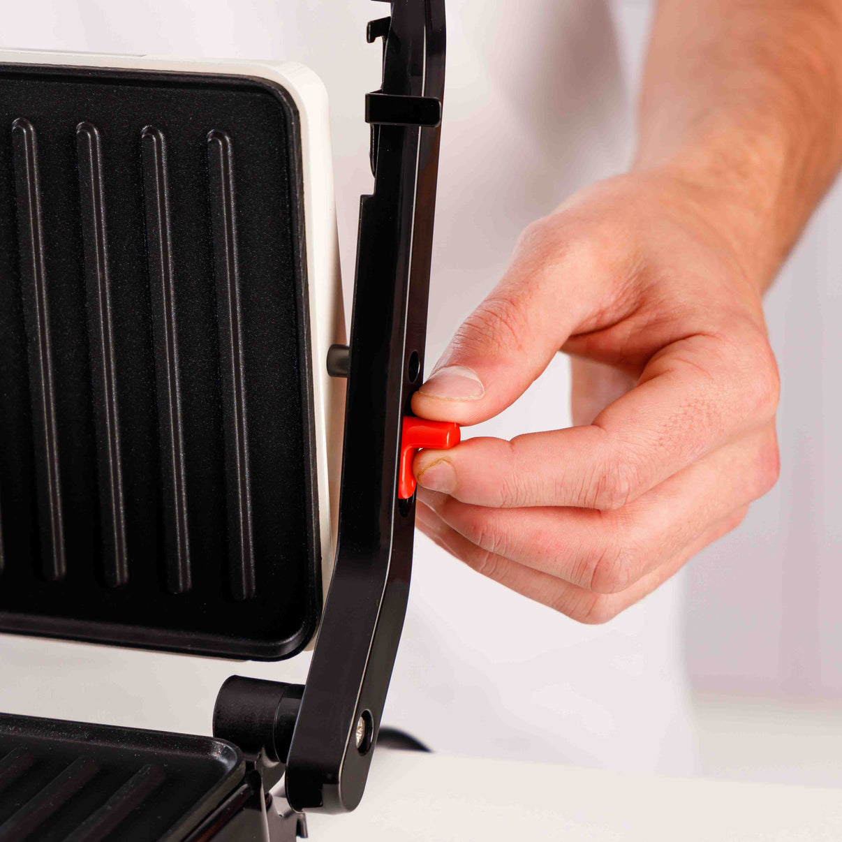 Una mano preme un pulsante rosso su una piastra per panini in bianco e nero. La piastra è aperta, mostrando le piastre della griglia.