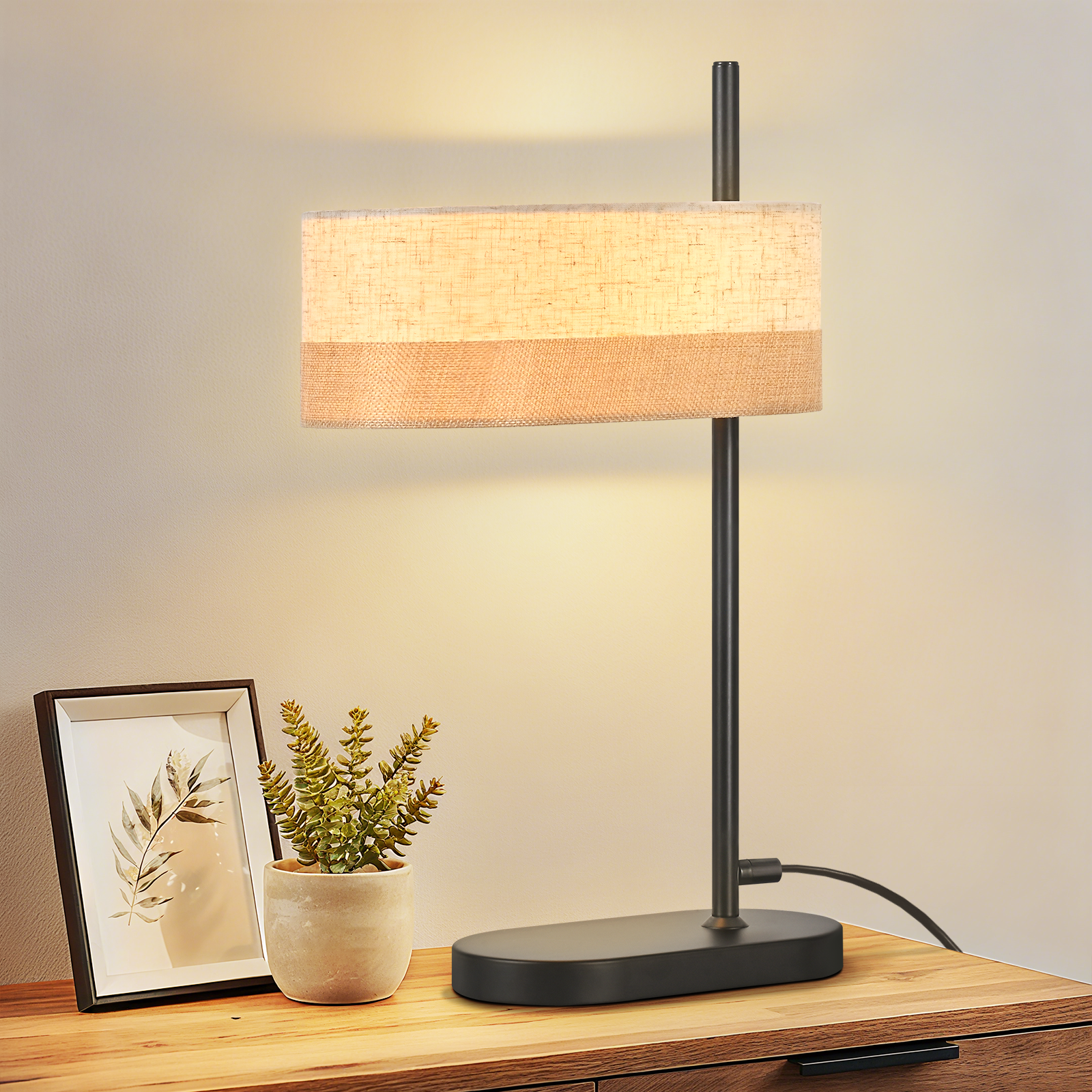 Lampa stołowa z prostokątnym abażurem stoi na drewnianej powierzchni. Obok znajduje się oprawiony obraz i roślina.