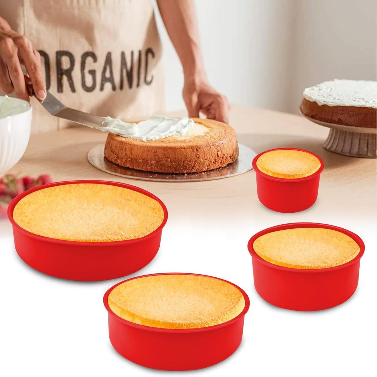 Eine Person, die einen Kuchen mit Zuckerguss verziert. Kuchenformen und gebackene Kuchen stehen auf einem Tisch. Schürze sagt 'ORGANIC'.